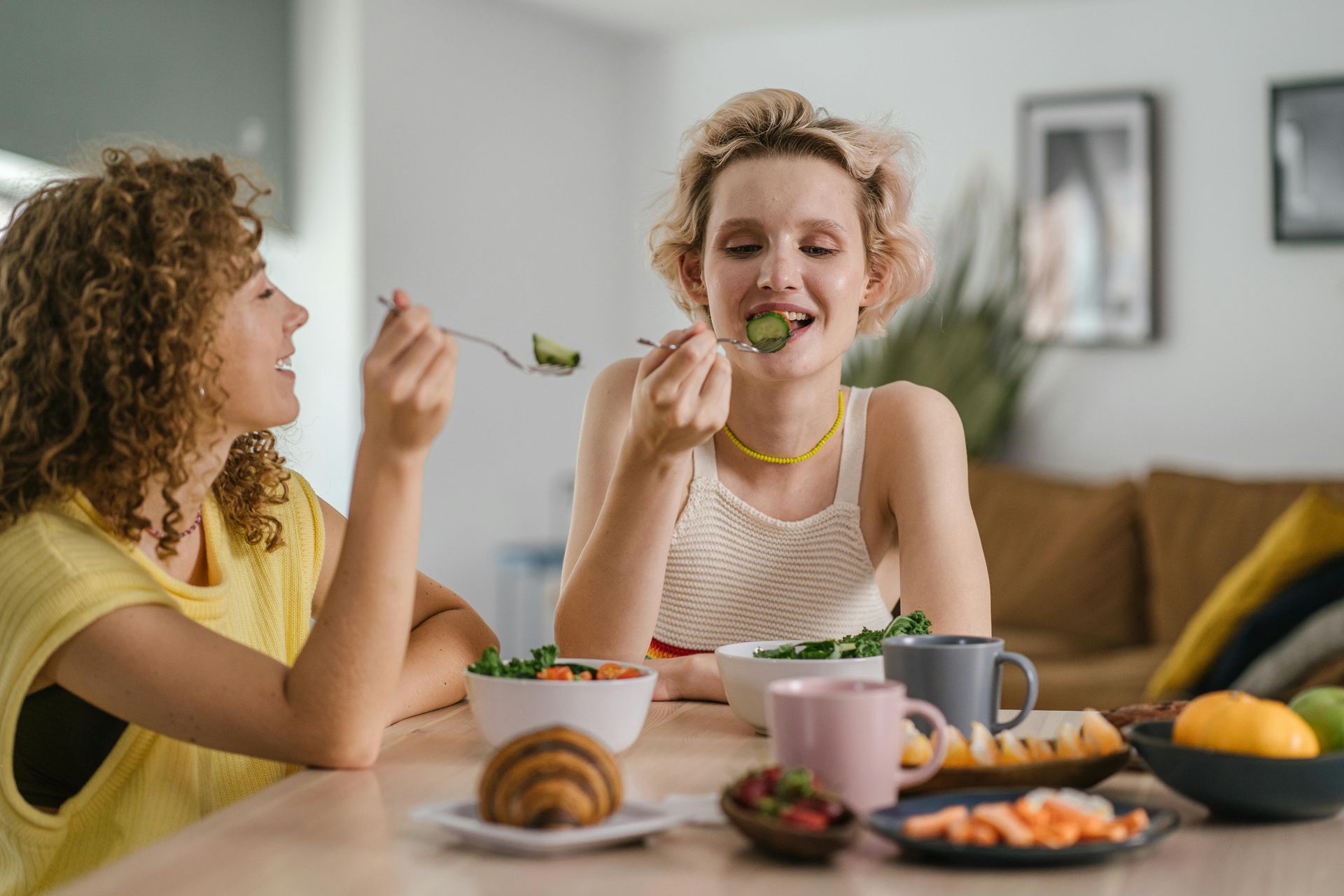 Two people sharing breakfast at a table, one eating a croissant in a bright living room.