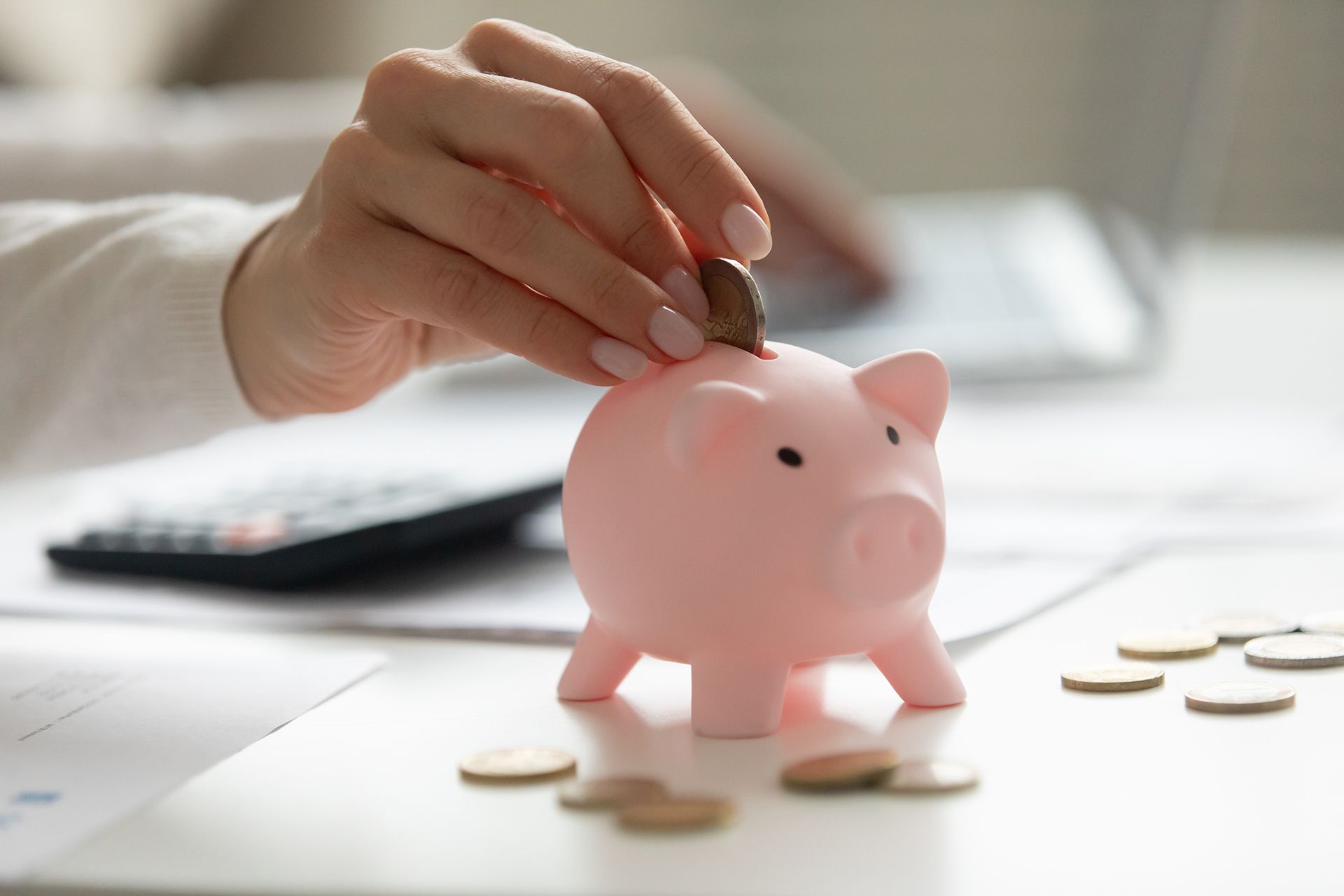 Hand placing a coin into a pink piggy bank on a white table with scattered coins