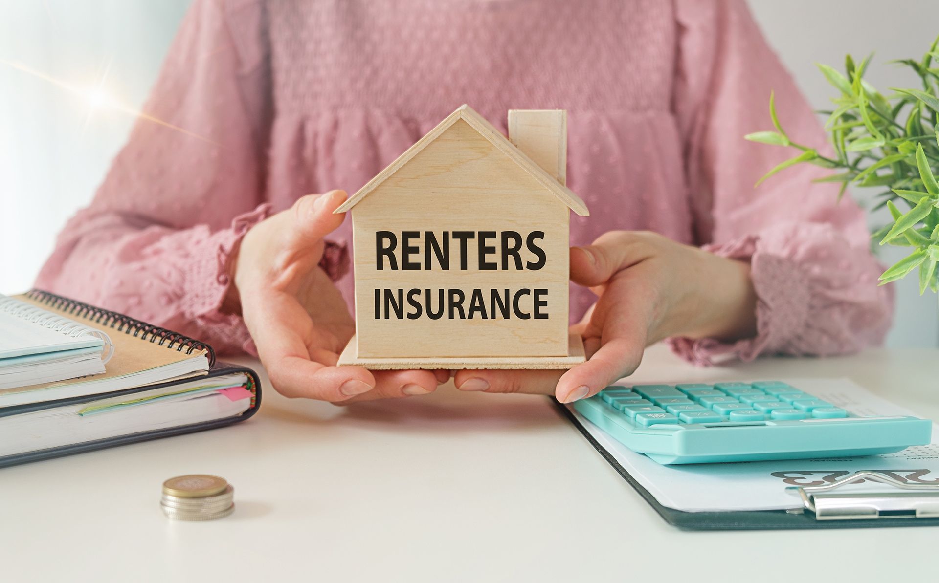 Hands holding a house model labeled “Renters Insurance” on a desk with notebooks and coins