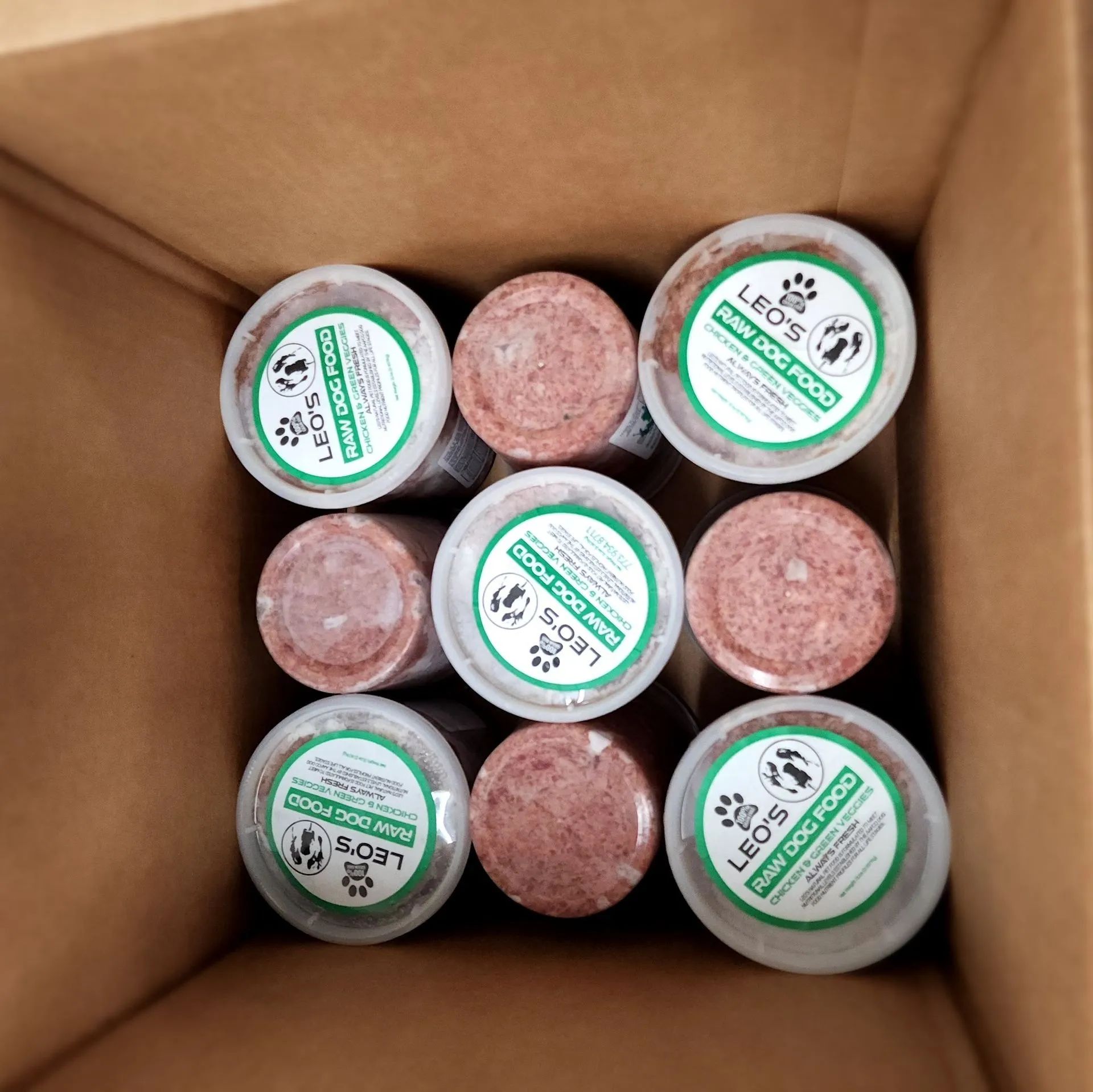 Open cardboard box holding six small jars with green labels and three pink round items