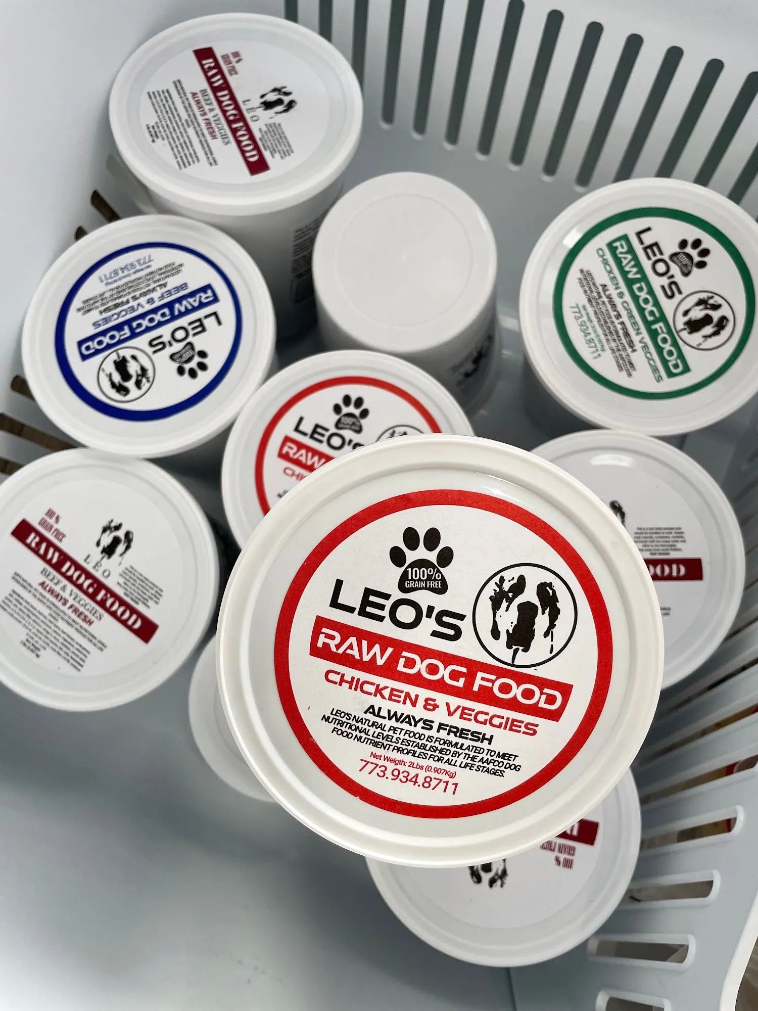 Various Leo’s raw dog food tubs in a plastic basket, with white lids and red and green labels