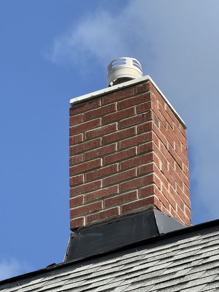 Brick chimney with a white vent cap on a shingled roof against a blue sky