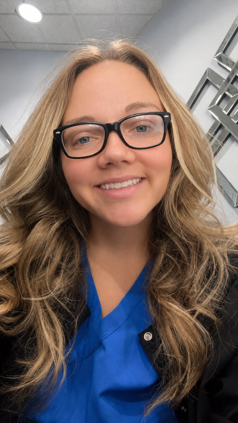Smiling person in blue scrubs and glasses, seated indoors with shelves in the background