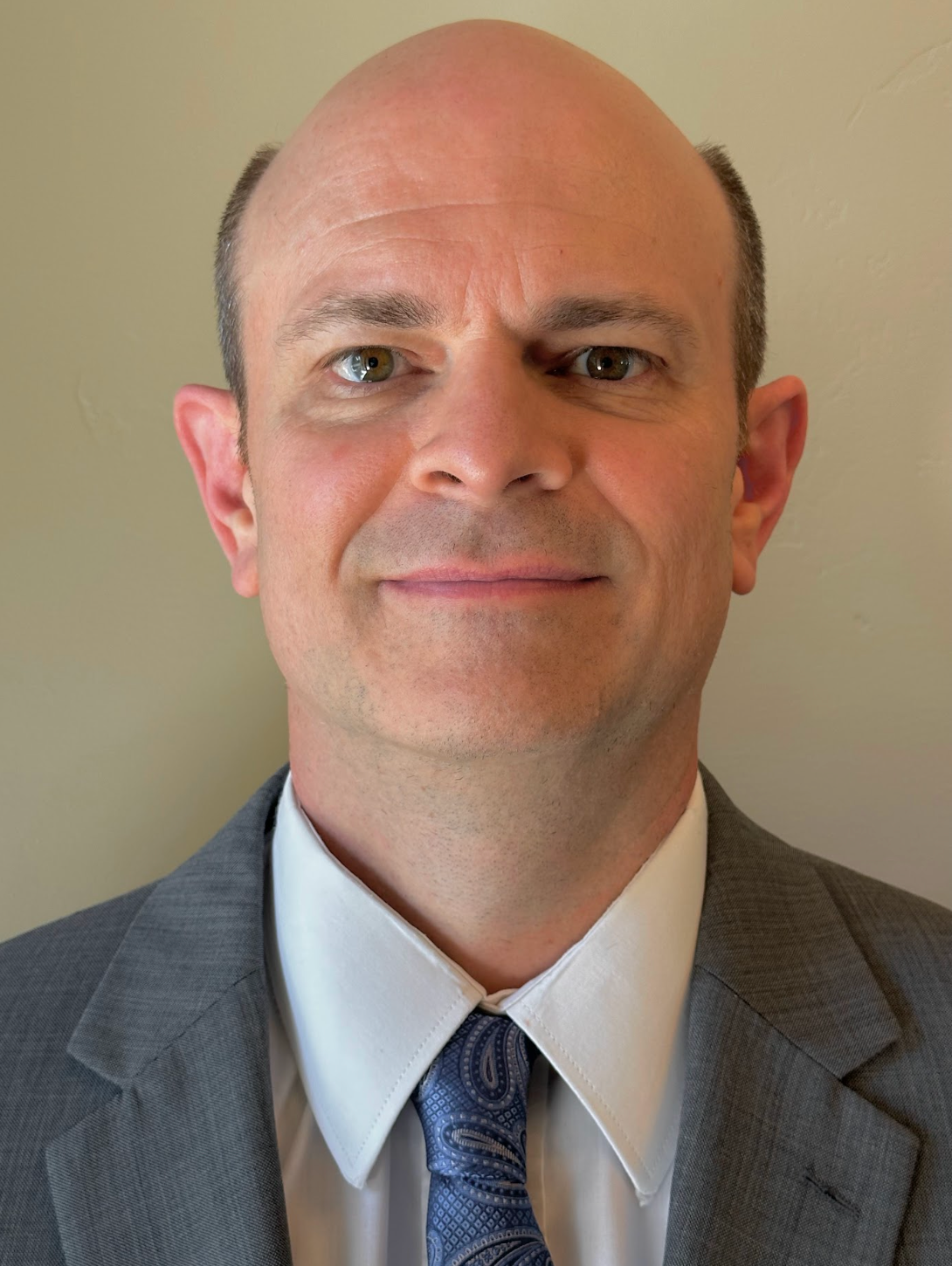 Portrait of a bald man in a gray suit and blue tie, facing the camera with a slight smile.
