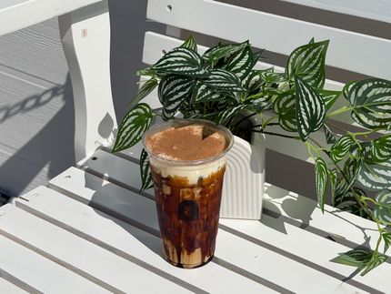 Iced coffee in a clear plastic cup on a white slatted table beside a striped potted plant.
