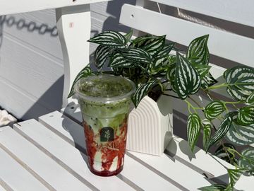 Pink strawberry drink in a clear cup beside a white chair and leafy plant in sunlight