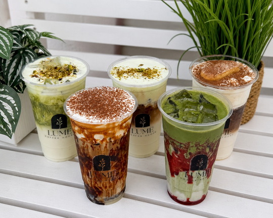 Five colorful iced drinks in clear cups on a white bench, topped with froth, herbs, and cocoa powder