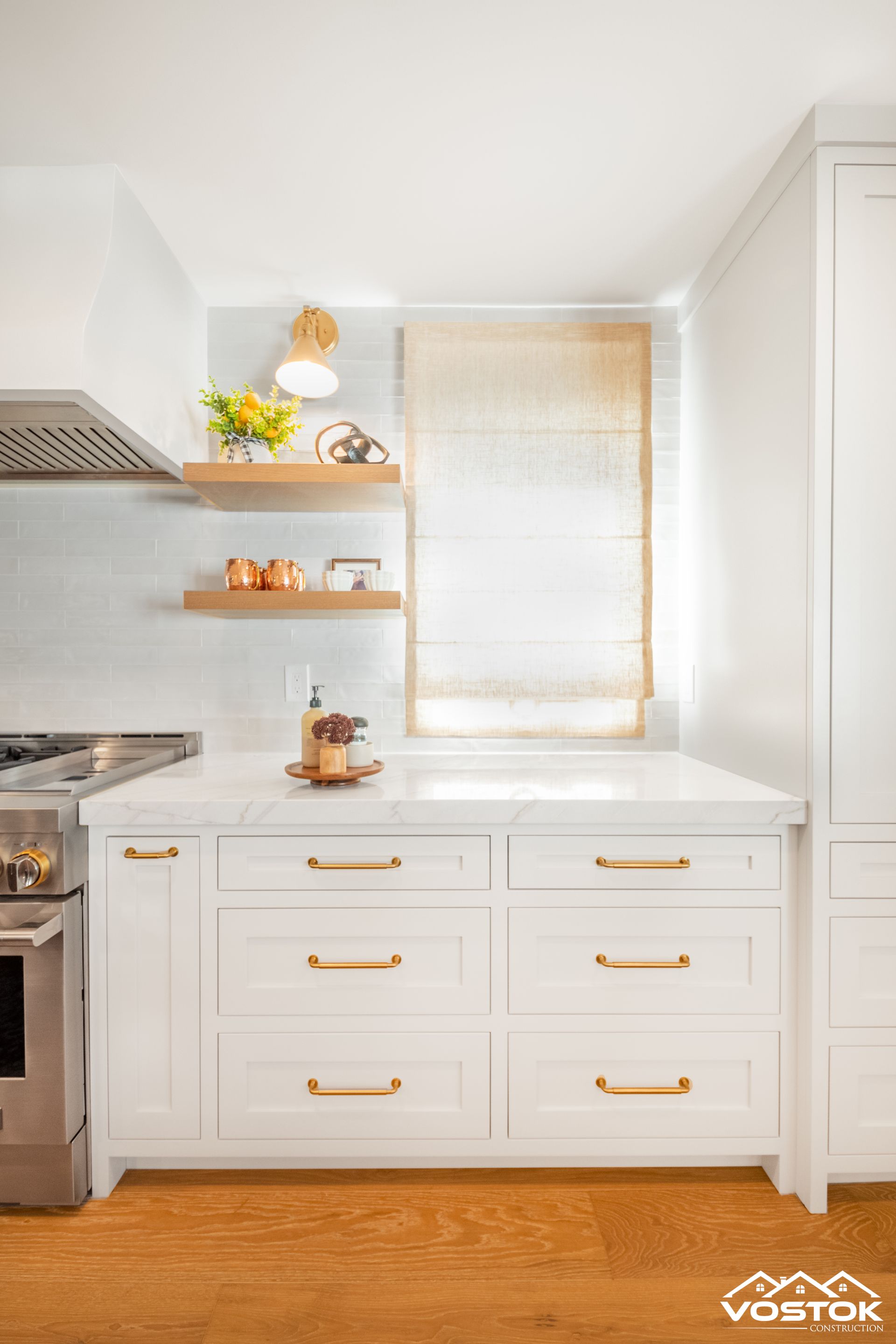 White kitchen cabinets with gold pulls, marble countertops, woven shade, and floating wood shelves