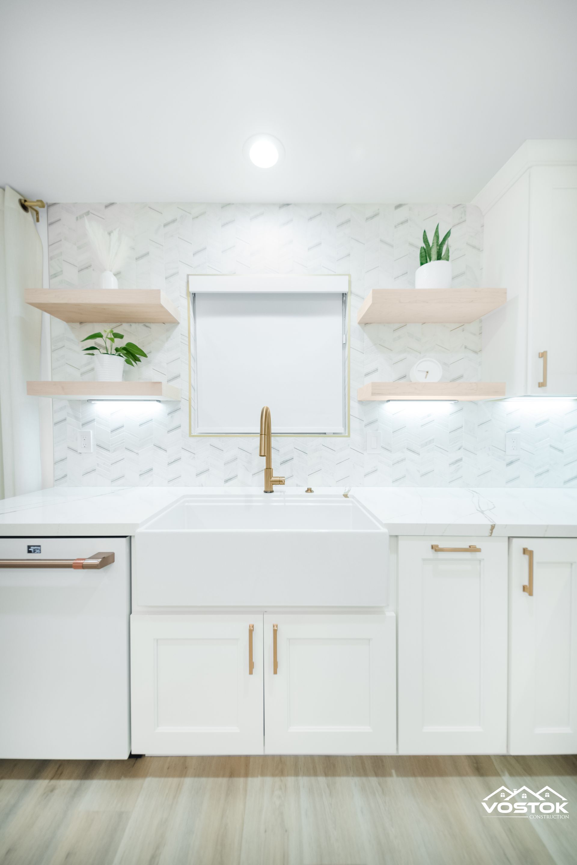 Bright white kitchen featuring farmhouse sink, gold faucet, herringbone backsplash, and floating wood shelves
