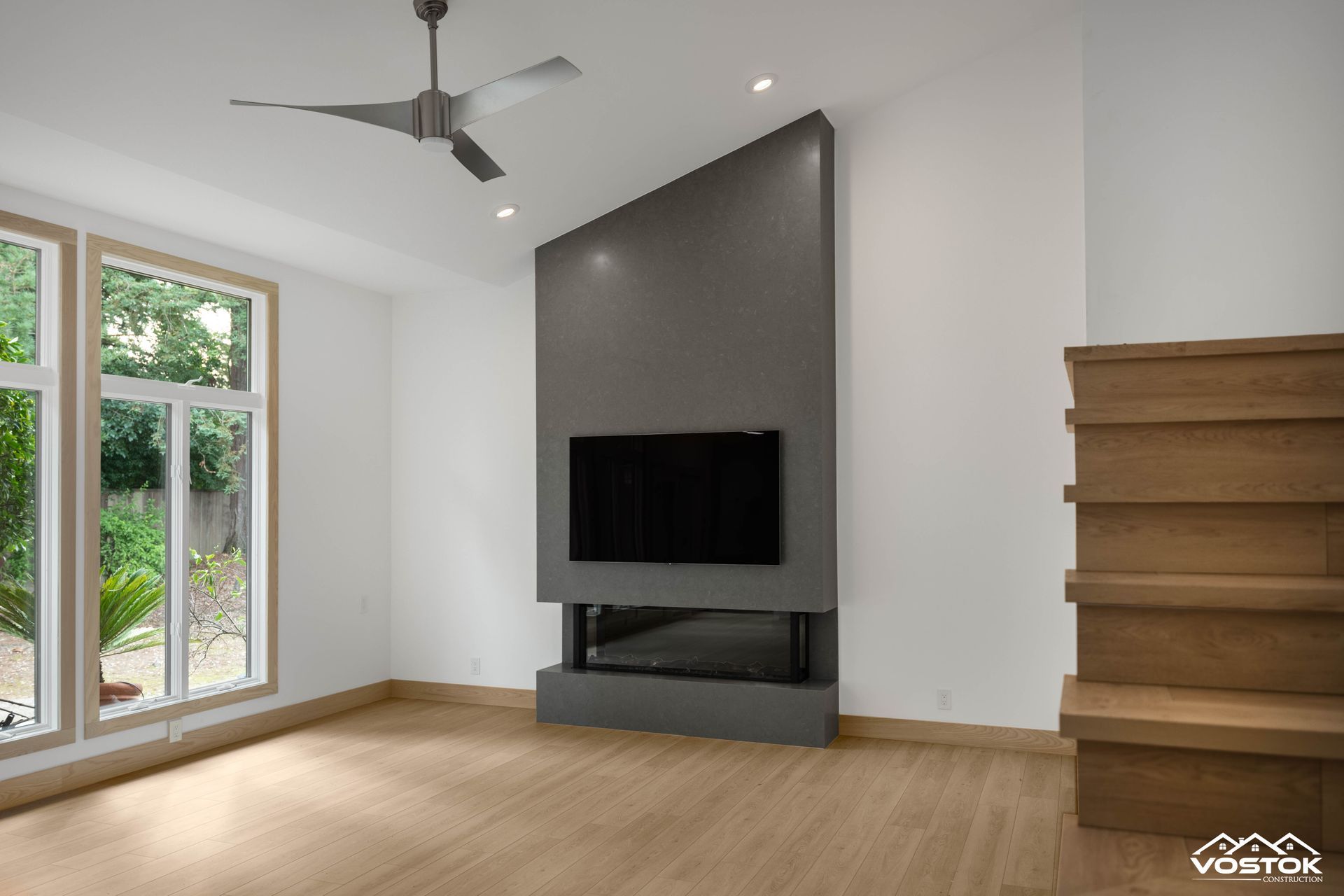 Modern living room with a prominent dark gray fireplace featuring an integrated TV, light wood flooring, white walls, and a contemporary silver ceiling fan.