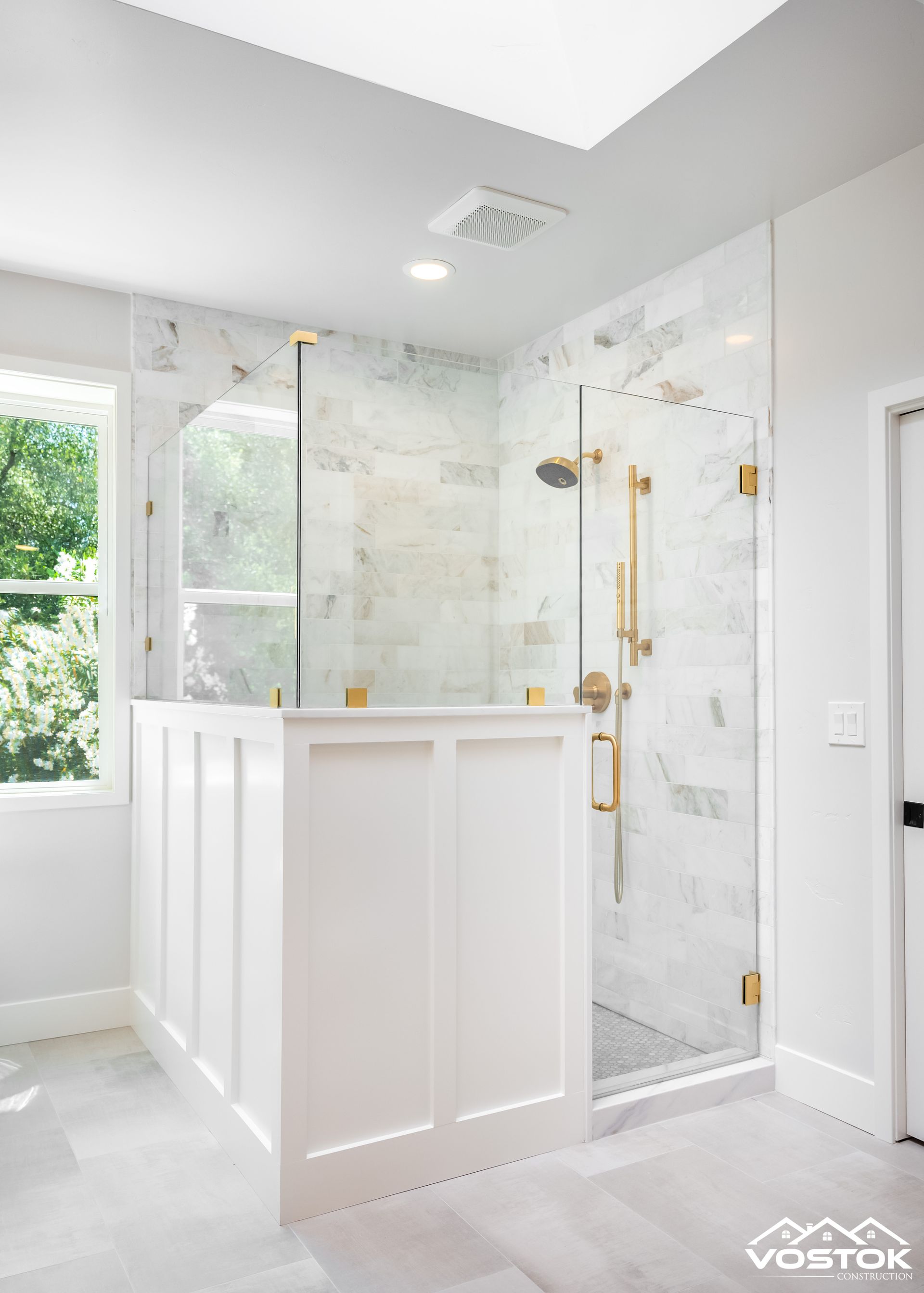 Luxurious walk-in shower with marble tile, gold hardware, glass door, and white wainscoting