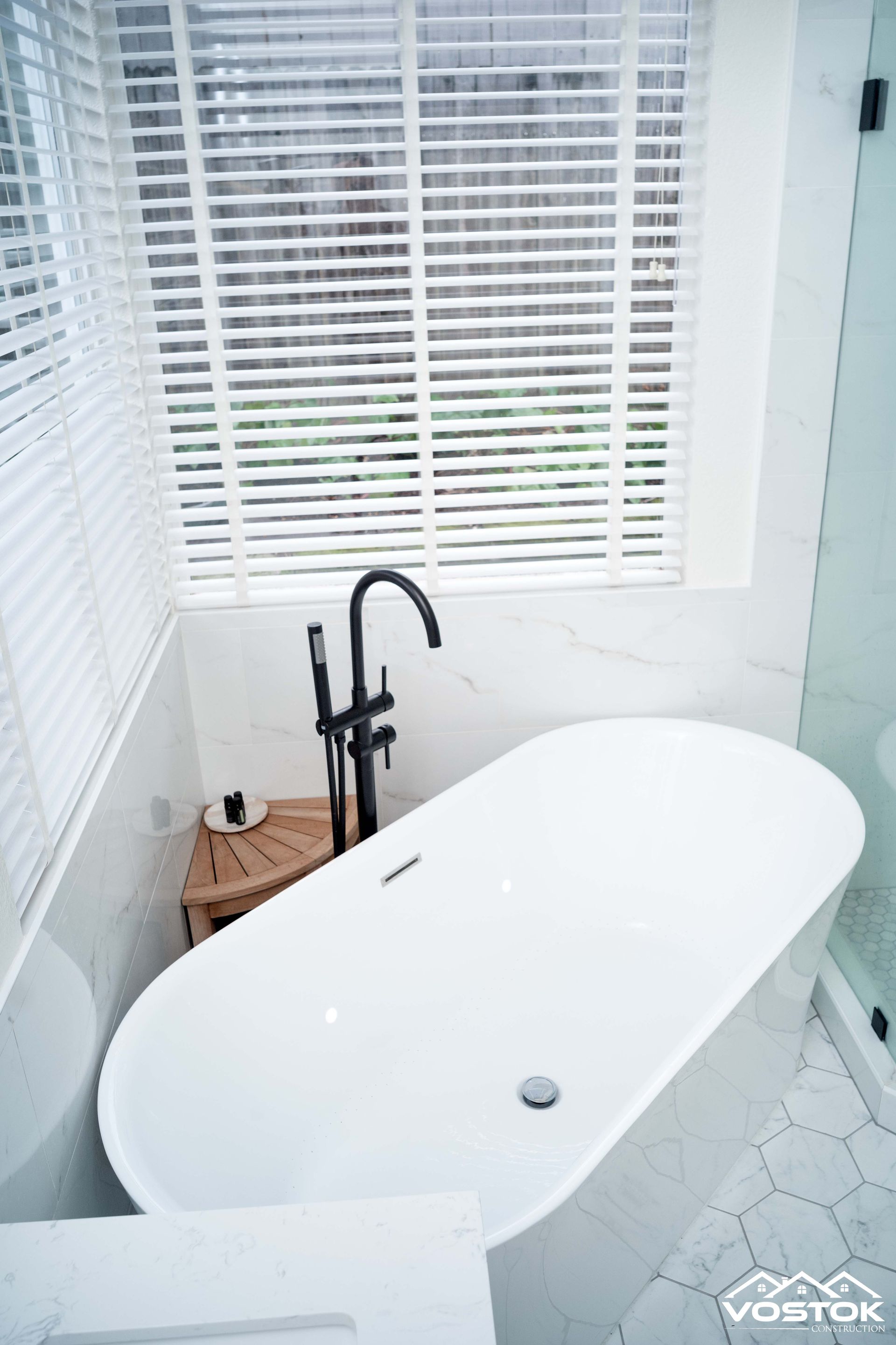 A white bathtub in a bathroom next to a window with blinds.