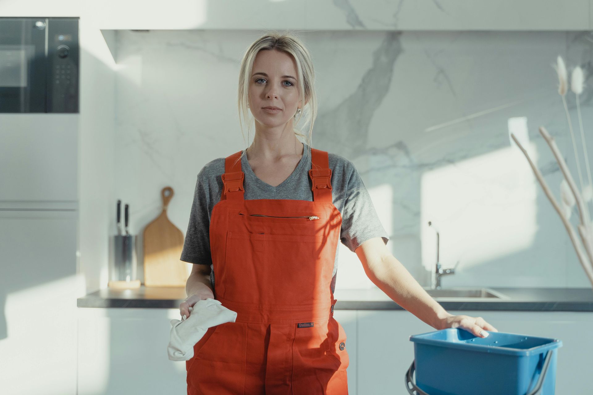 A woman in orange overalls is standing in a kitchen holding a mop and bucket.