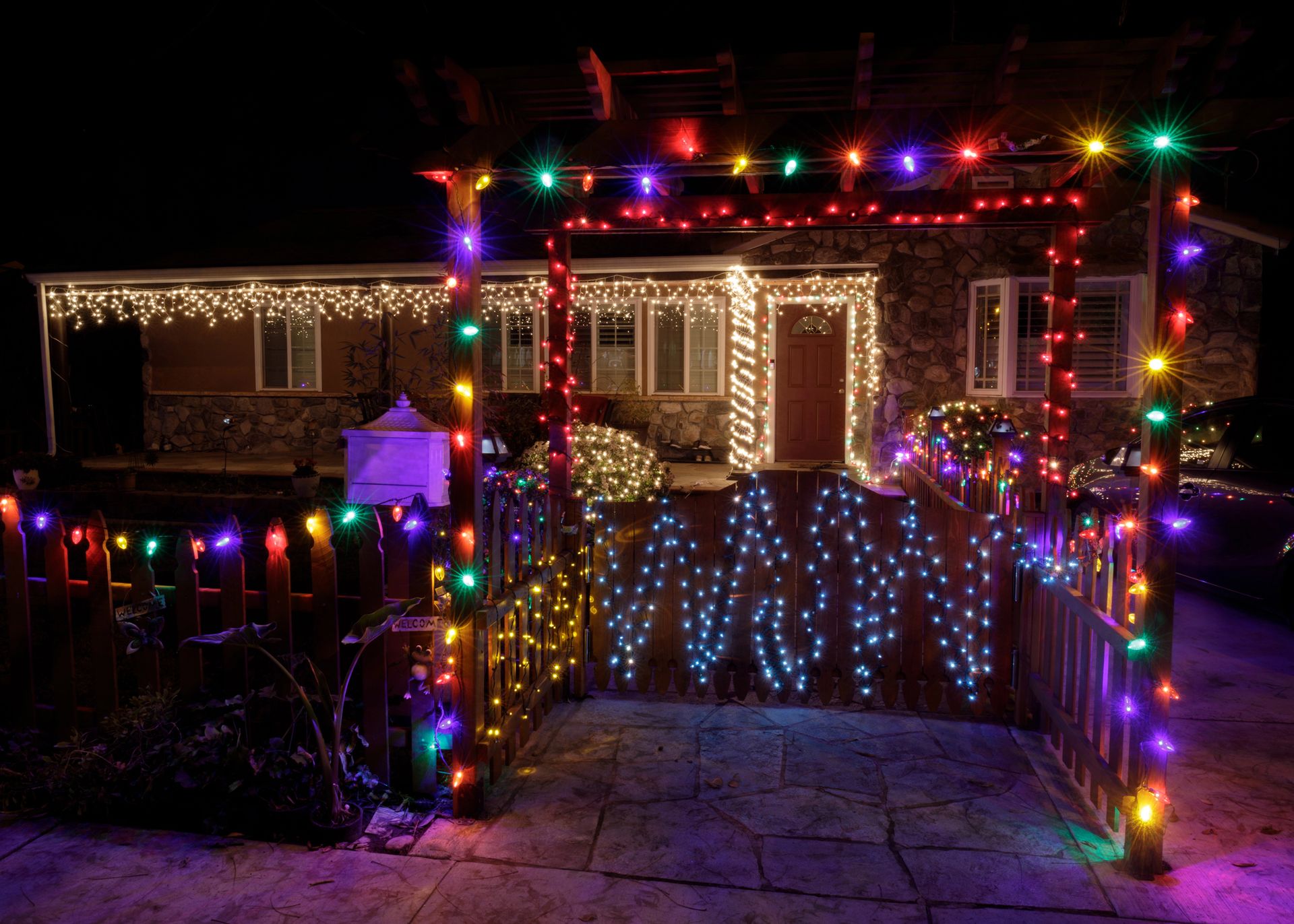 House and porch lit with colorful Christmas lights at night, with a glowing front gate and walkway
