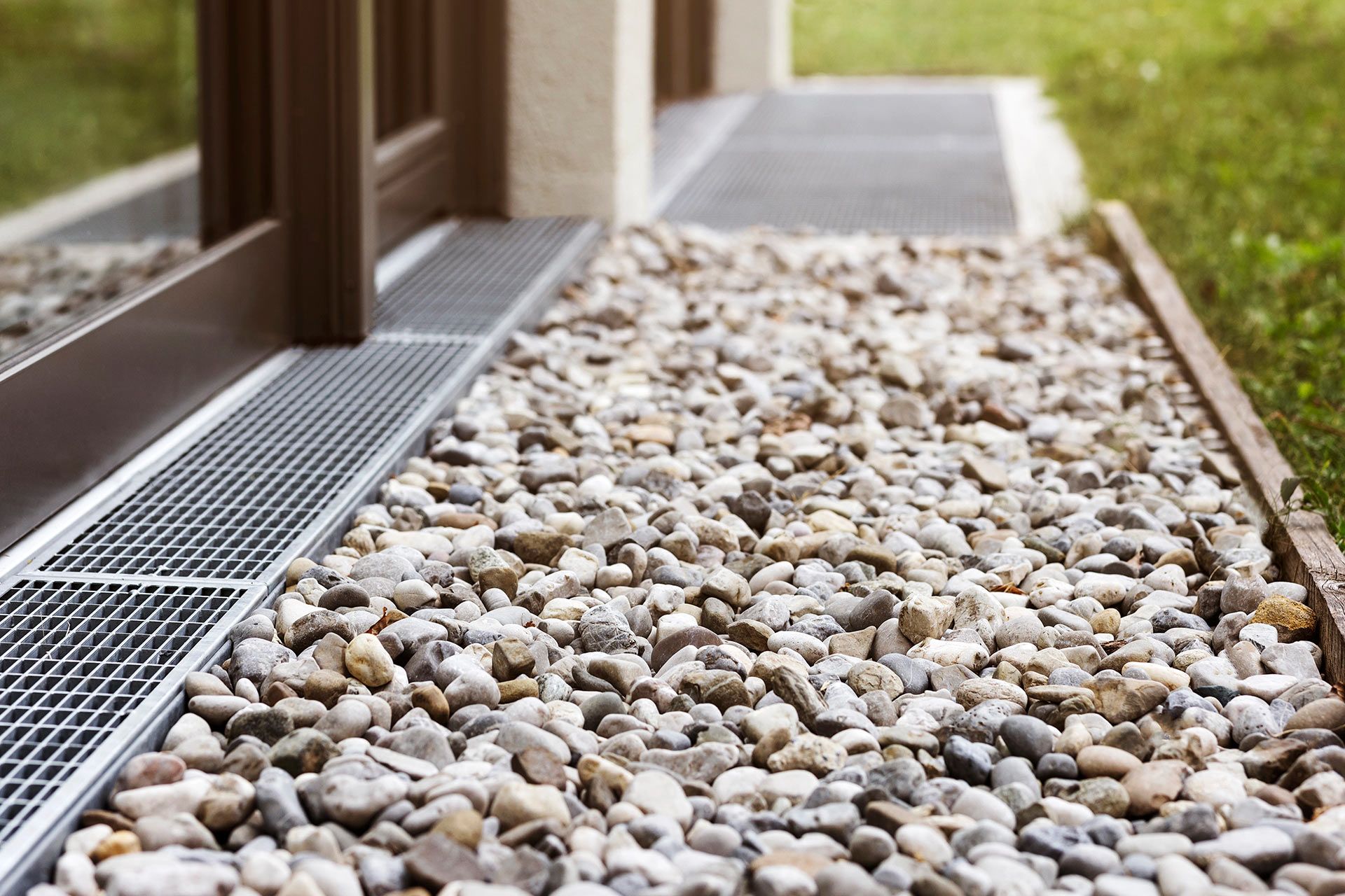 Gravel-covered walkway alongside a building, with a metal drainage grate near a window.