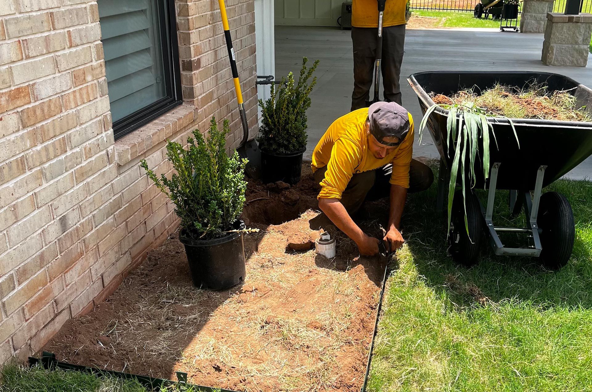 Landscaper installing edging next to a house; a wheelbarrow of debris is nearby.