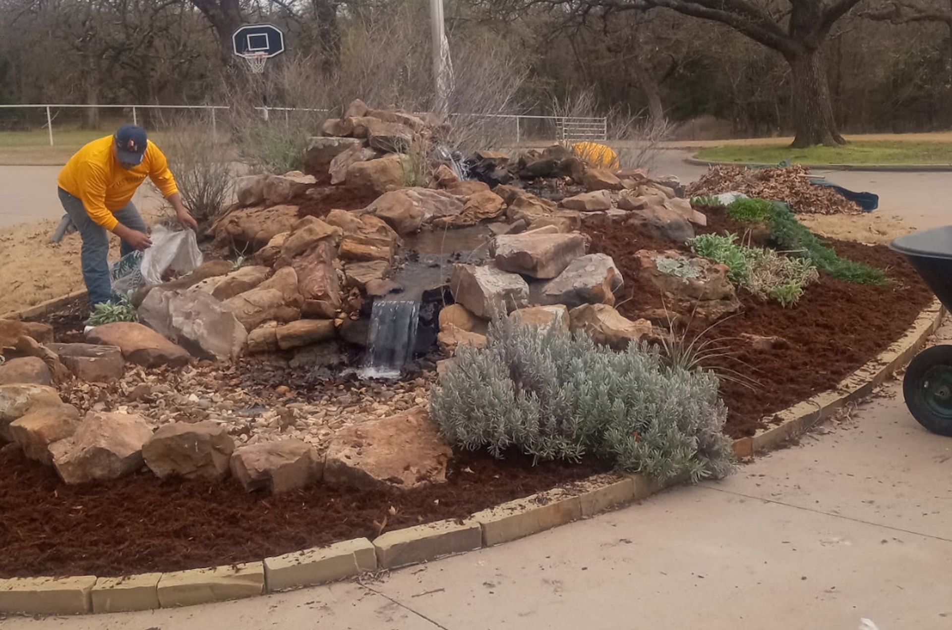 Man in yellow jacket adding something to a rock garden with a small waterfall.