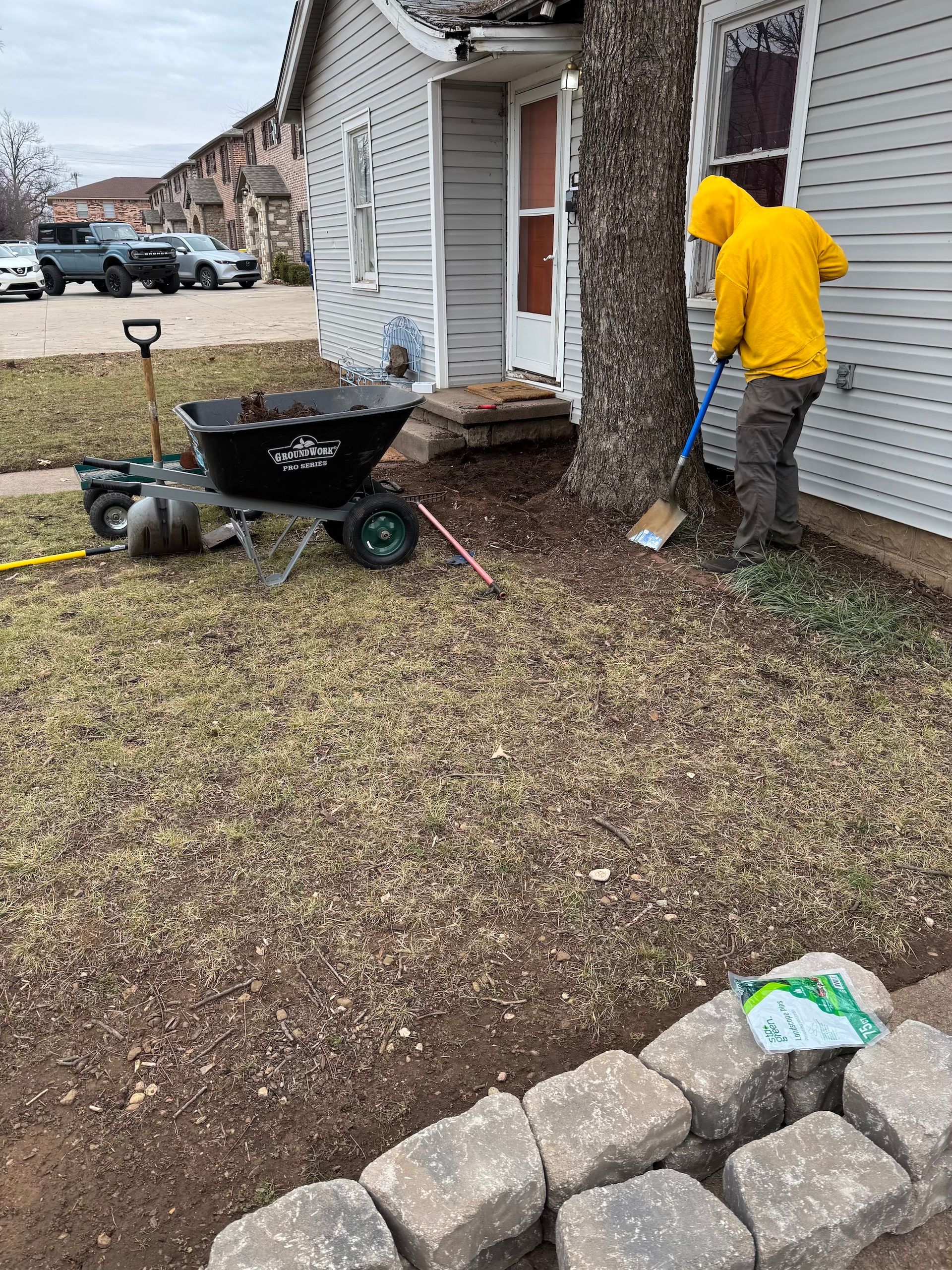 Man in yellow jacket mulching around a tree next to a house. A wheelbarrow and tools sit nearby.