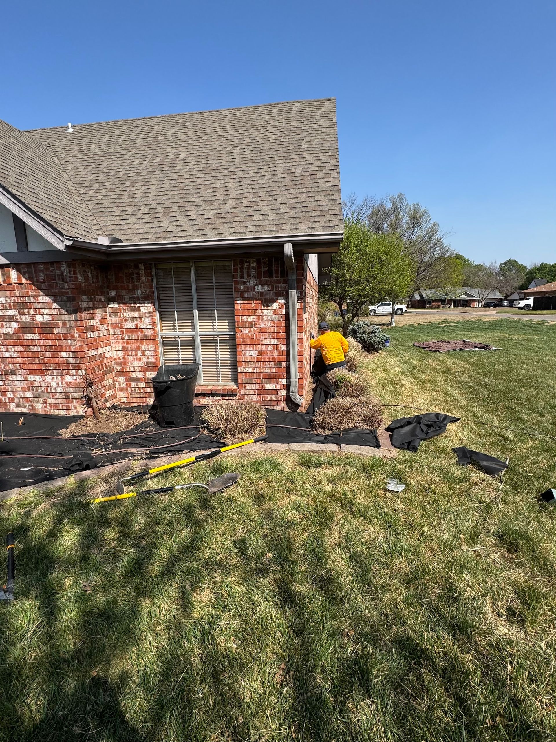 Person in yellow shirt working on landscaping next to brick house.