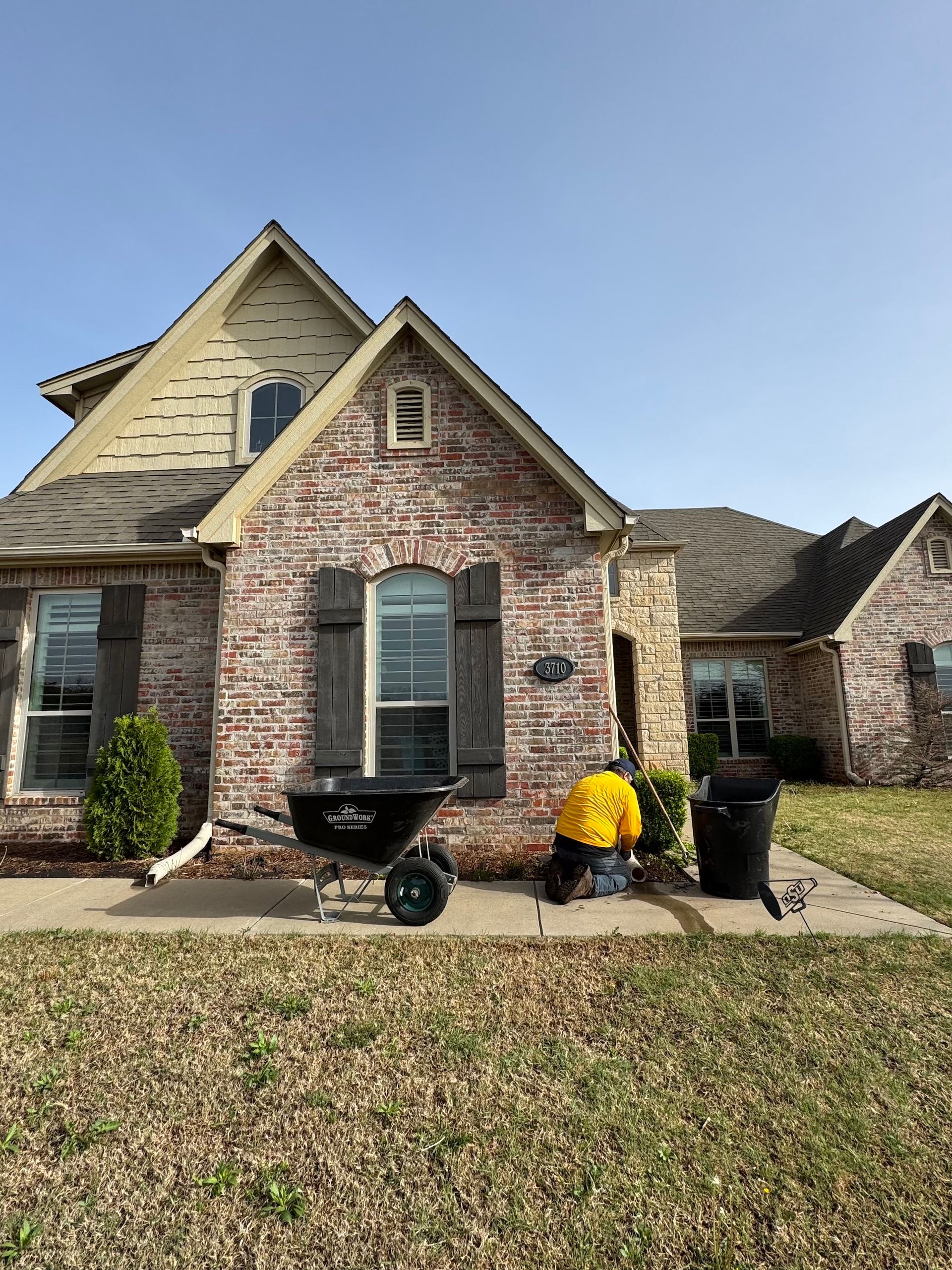 Person kneeling near a house, likely working on the landscaping. Wheelbarrow and trash can nearby.
