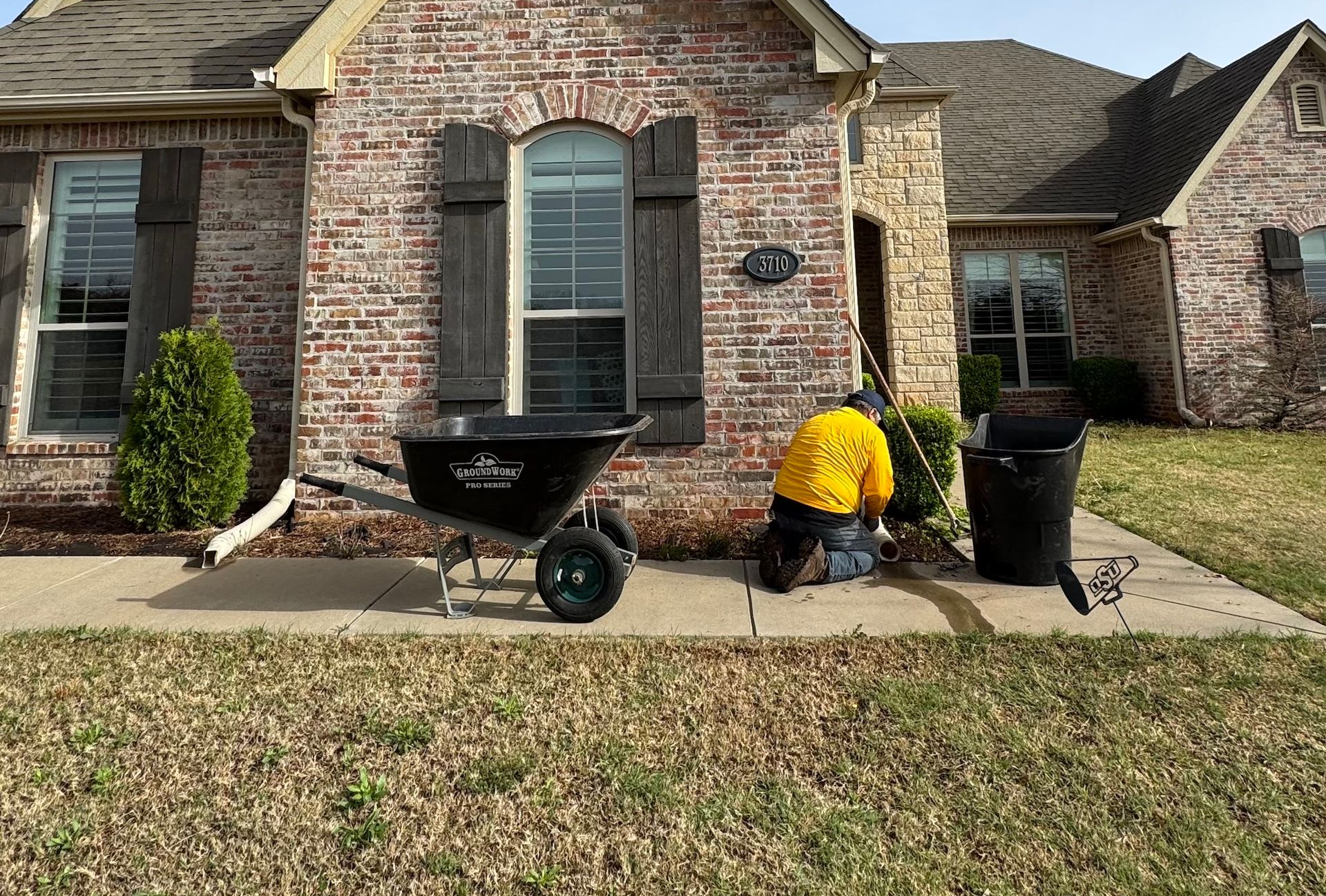 Person kneeling by a house trimming bushes; a wheelbarrow and trash can are nearby.
