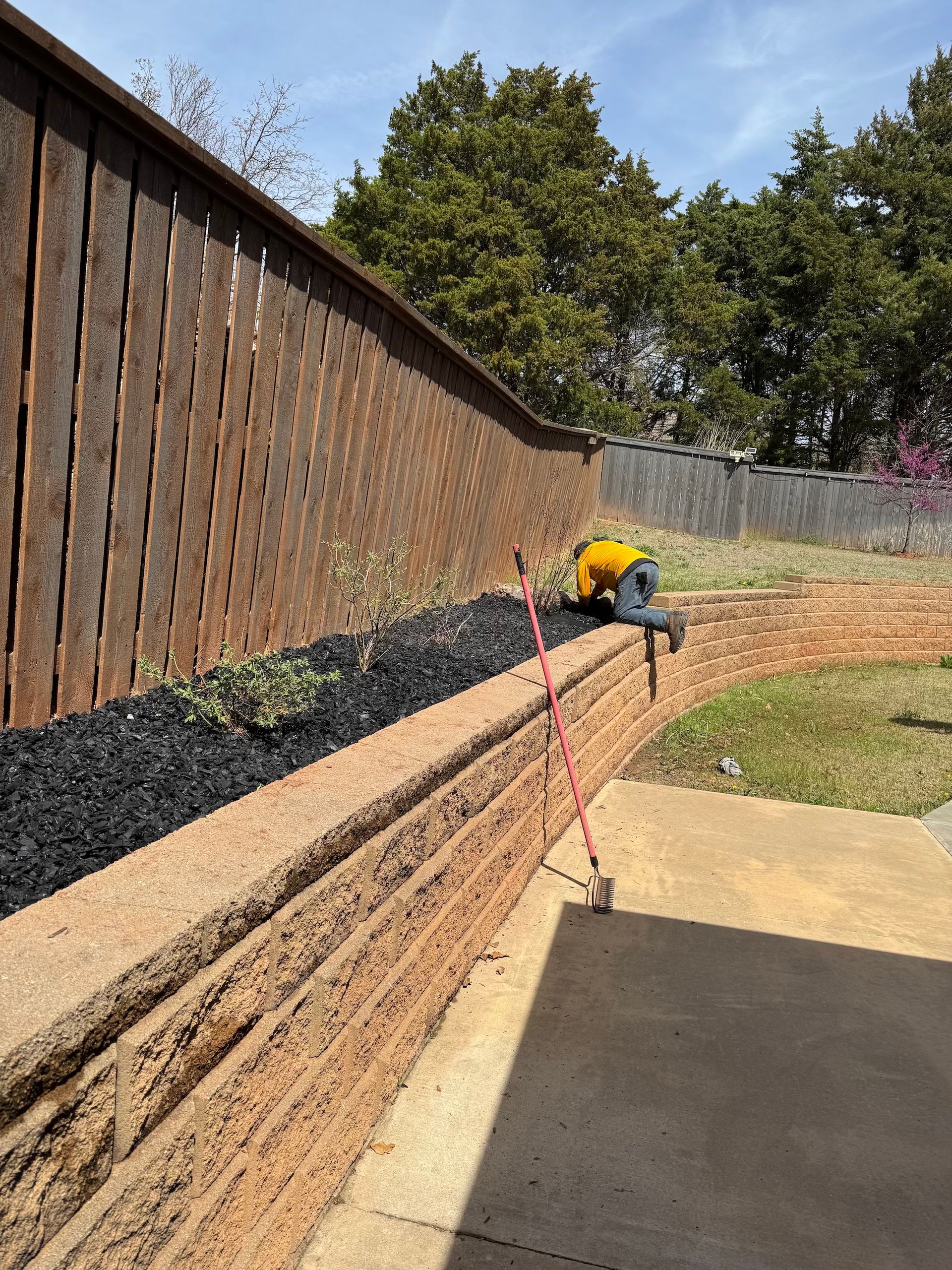Person working on landscaping near a brown fence and retaining wall; black mulch and a pink rake.