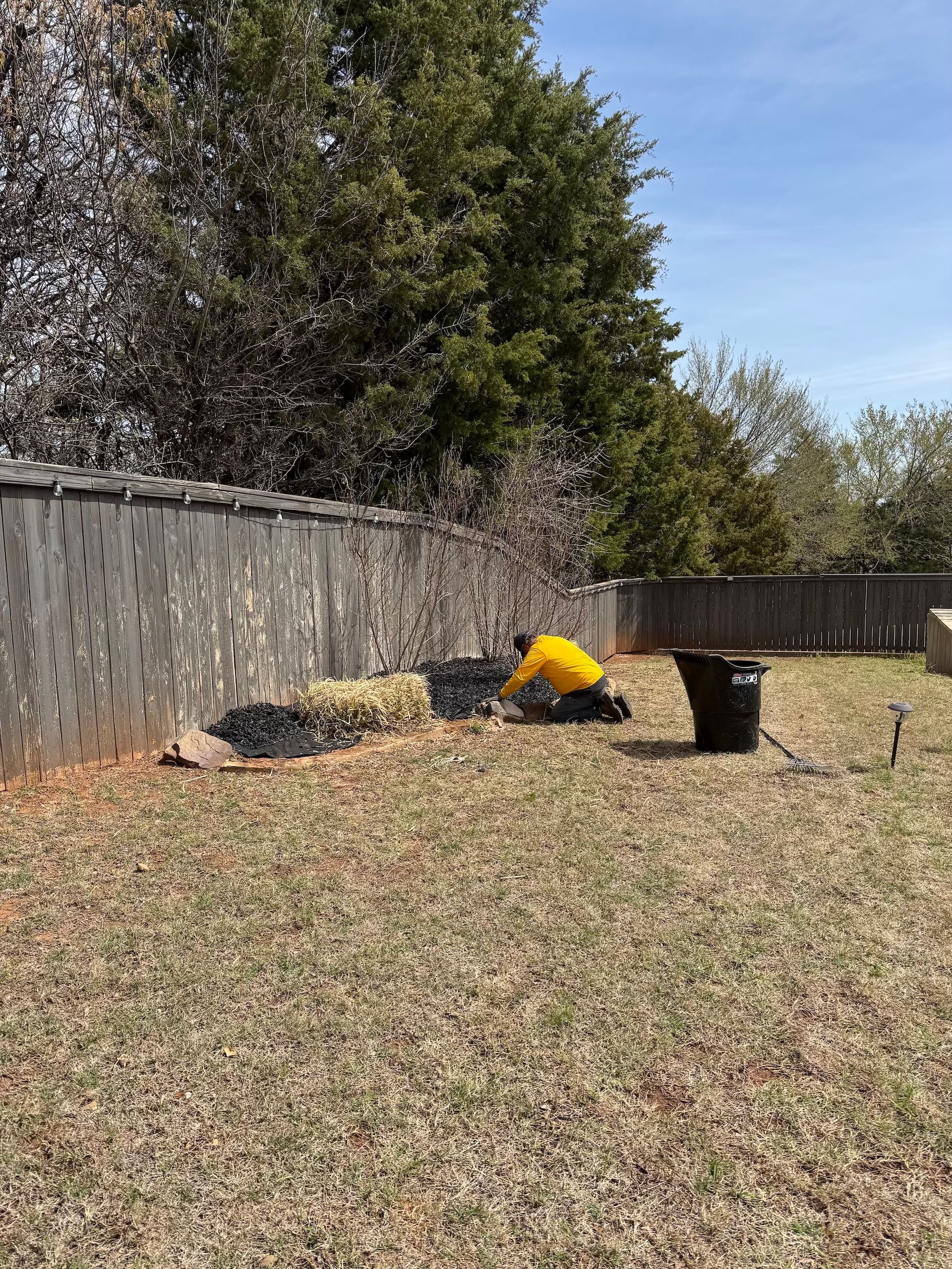 Person in yellow shirt kneeling, working in garden bed next to a wooden fence.