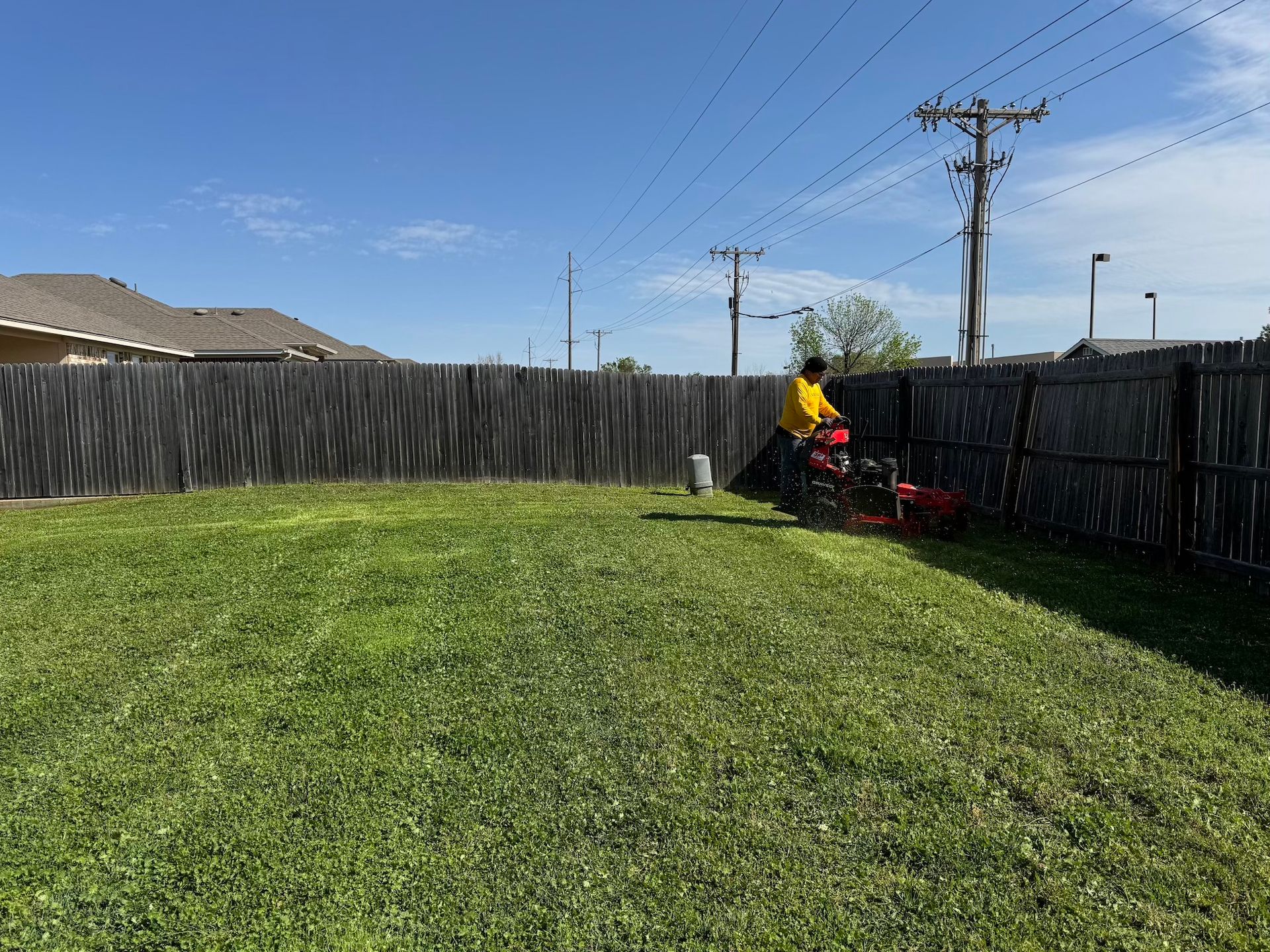 Person mowing a green lawn with a red mower in a backyard with a wooden fence and clear blue sky.
