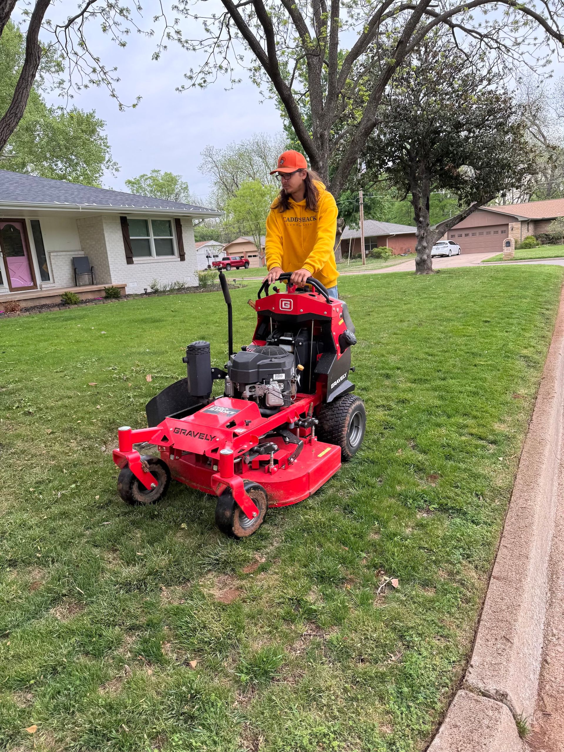 Person in yellow jacket mowing a lawn with a red zero-turn mower in a residential area.