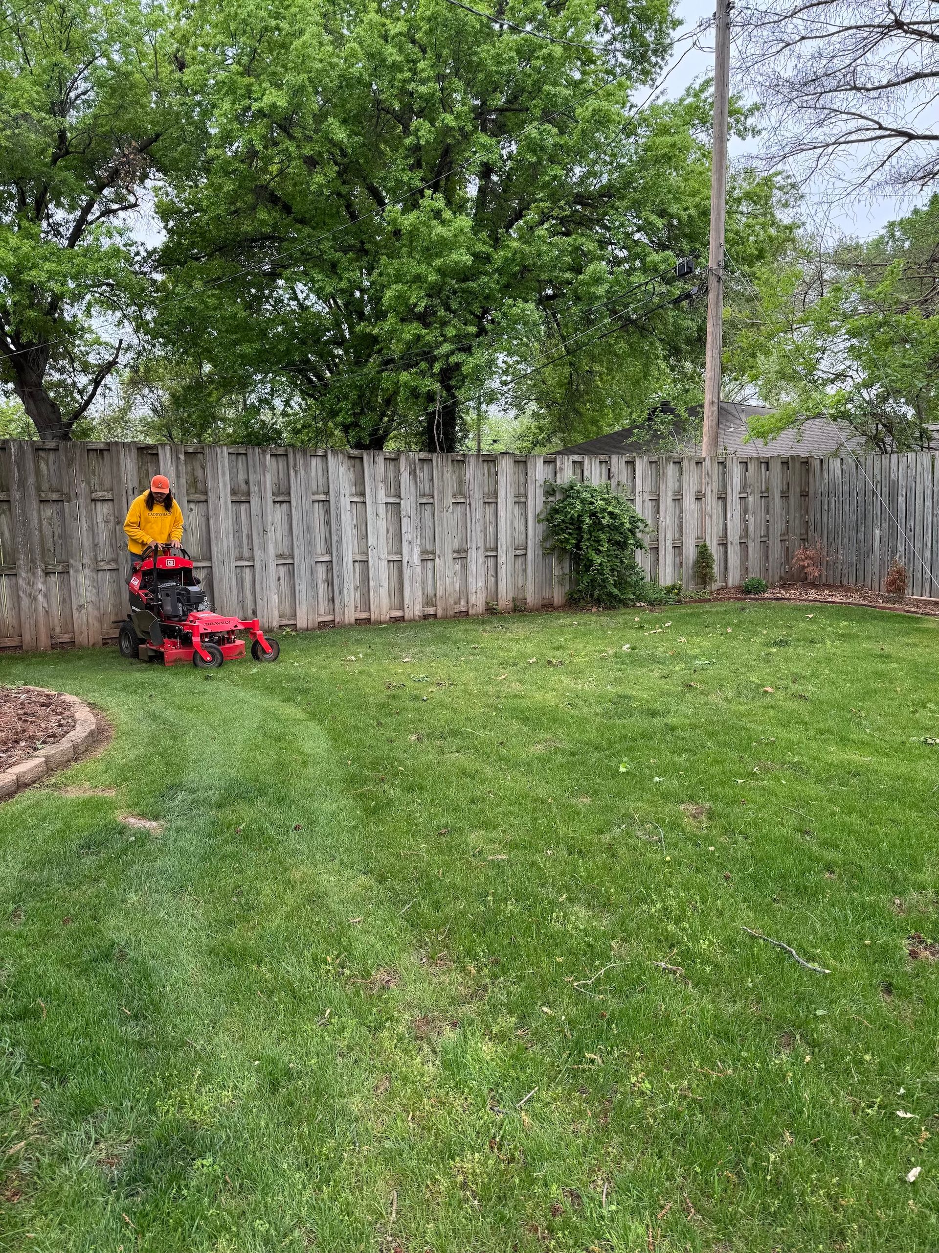 Person mowing a green lawn with a red lawnmower in a backyard with a wooden fence.