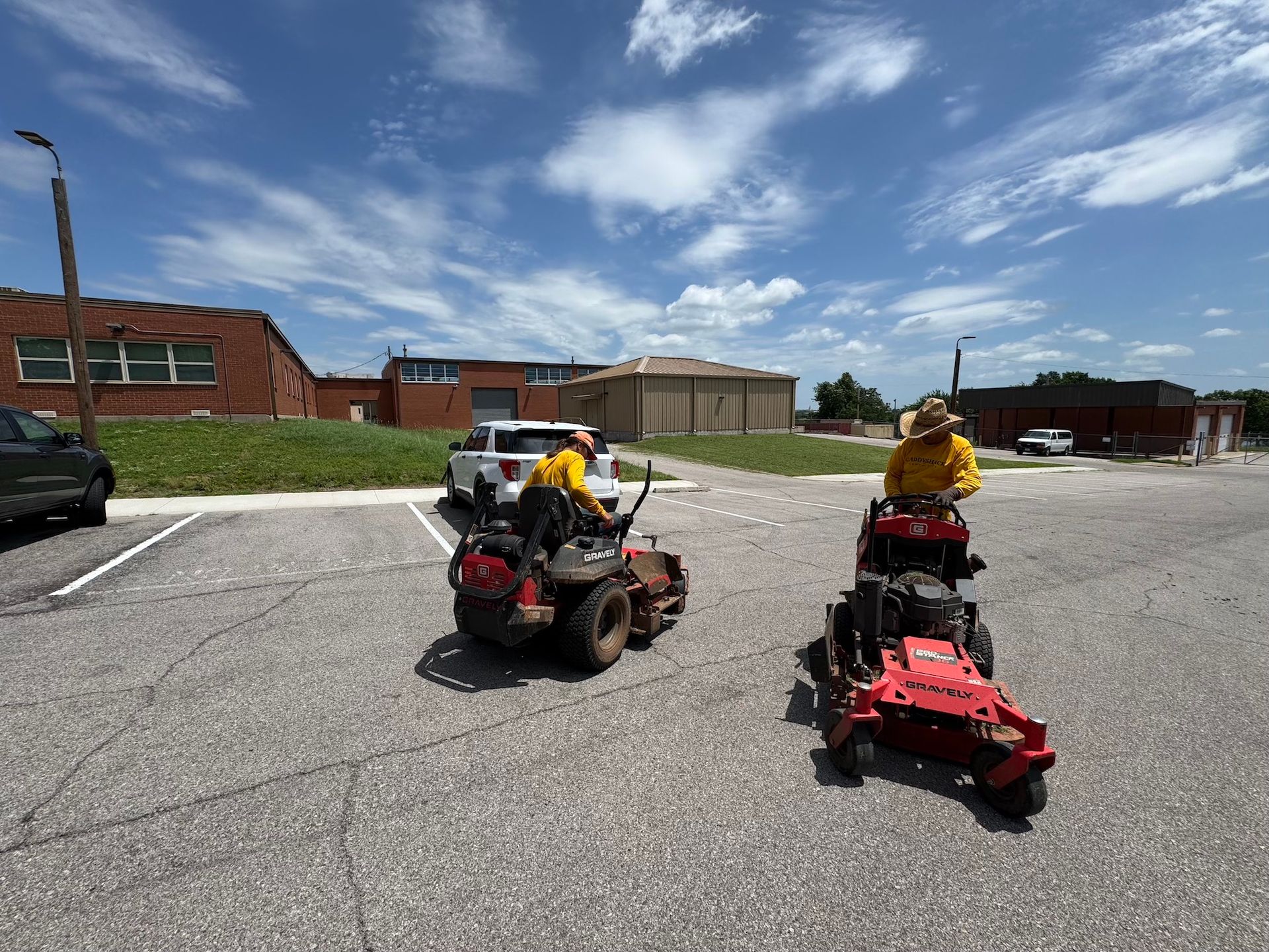Two people on riding lawnmowers in a parking lot. Sunny, blue sky. Buildings in the background.
