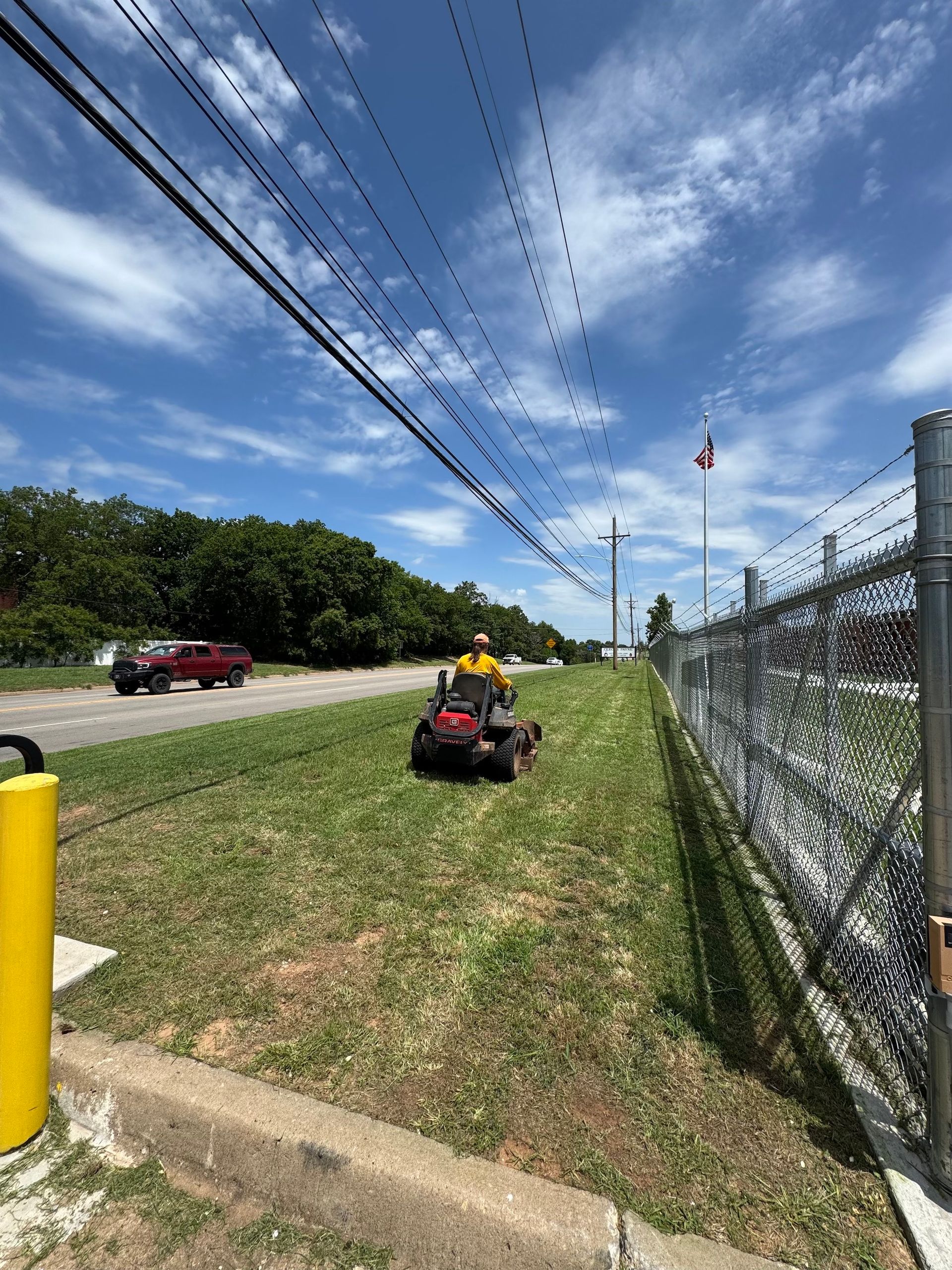 Person mowing grass alongside a chain-link fence and road on a sunny day.