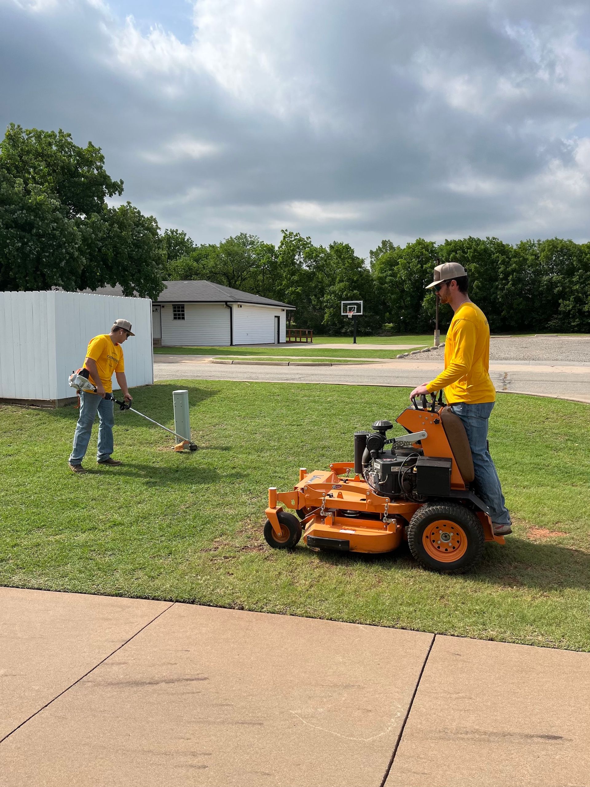 Two men mowing lawn on a cloudy day. One uses a weed eater, the other rides a yellow lawnmower.