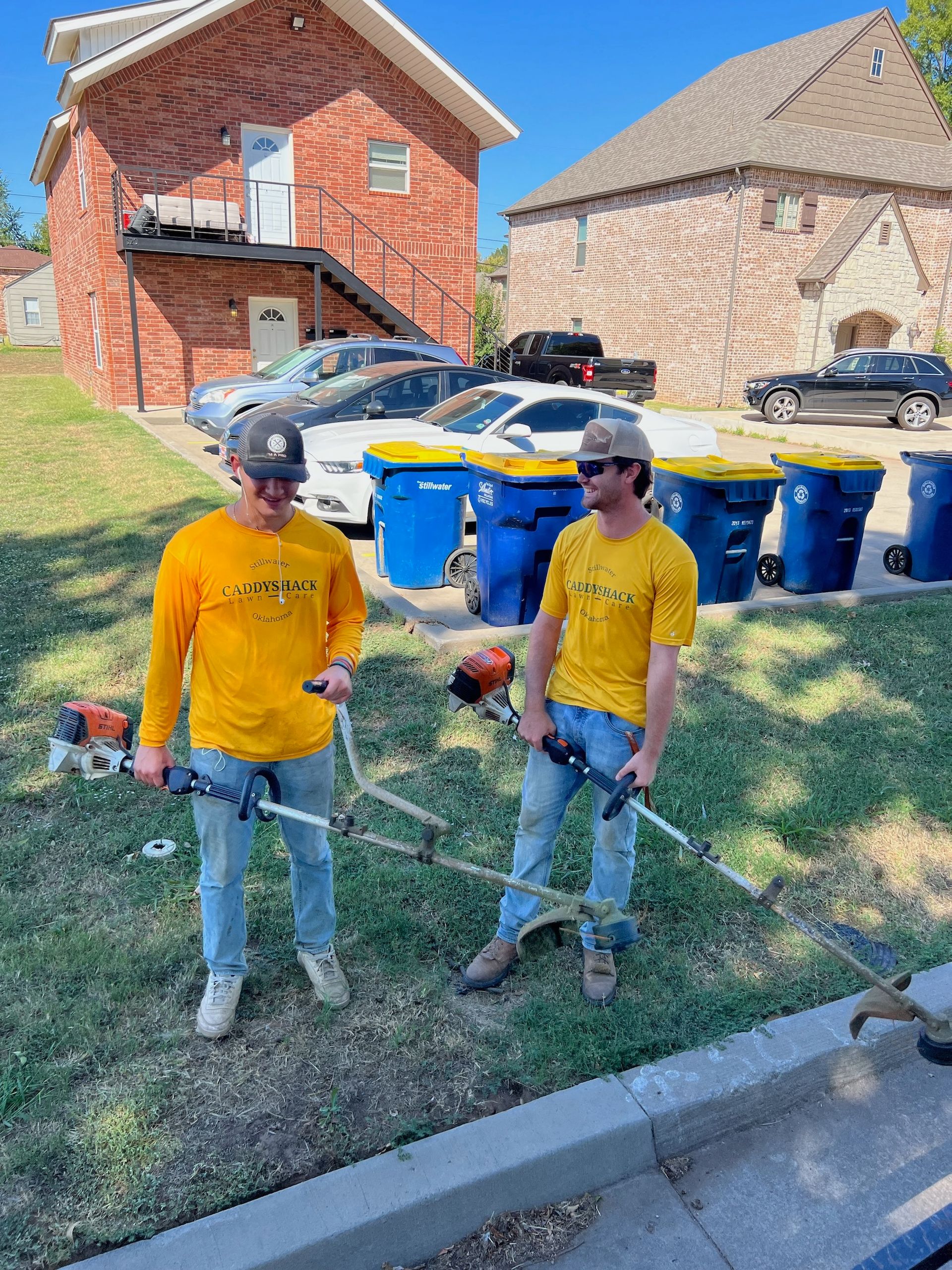 Two men in yellow shirts holding weed eaters, on a grassy lawn, near a street and buildings.