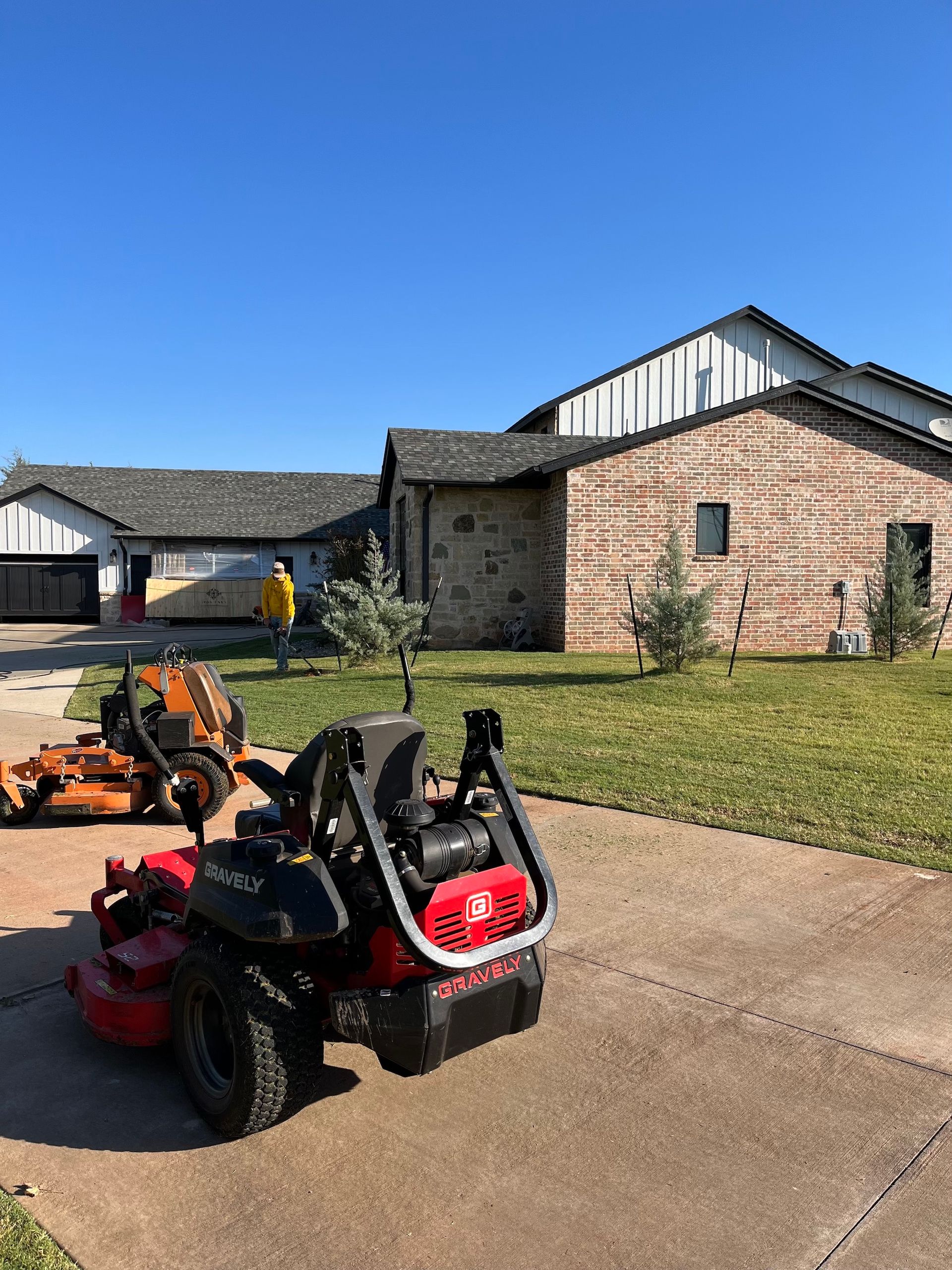 Lawn mowers parked on driveway in front of a brick house with a blue sky. A person stands on the lawn.