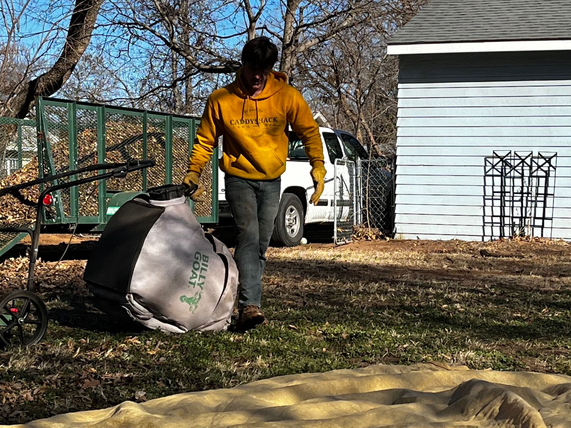 Man in yellow hoodie carrying a full lawn bag in a yard, with a mower nearby and a blue house in the background.