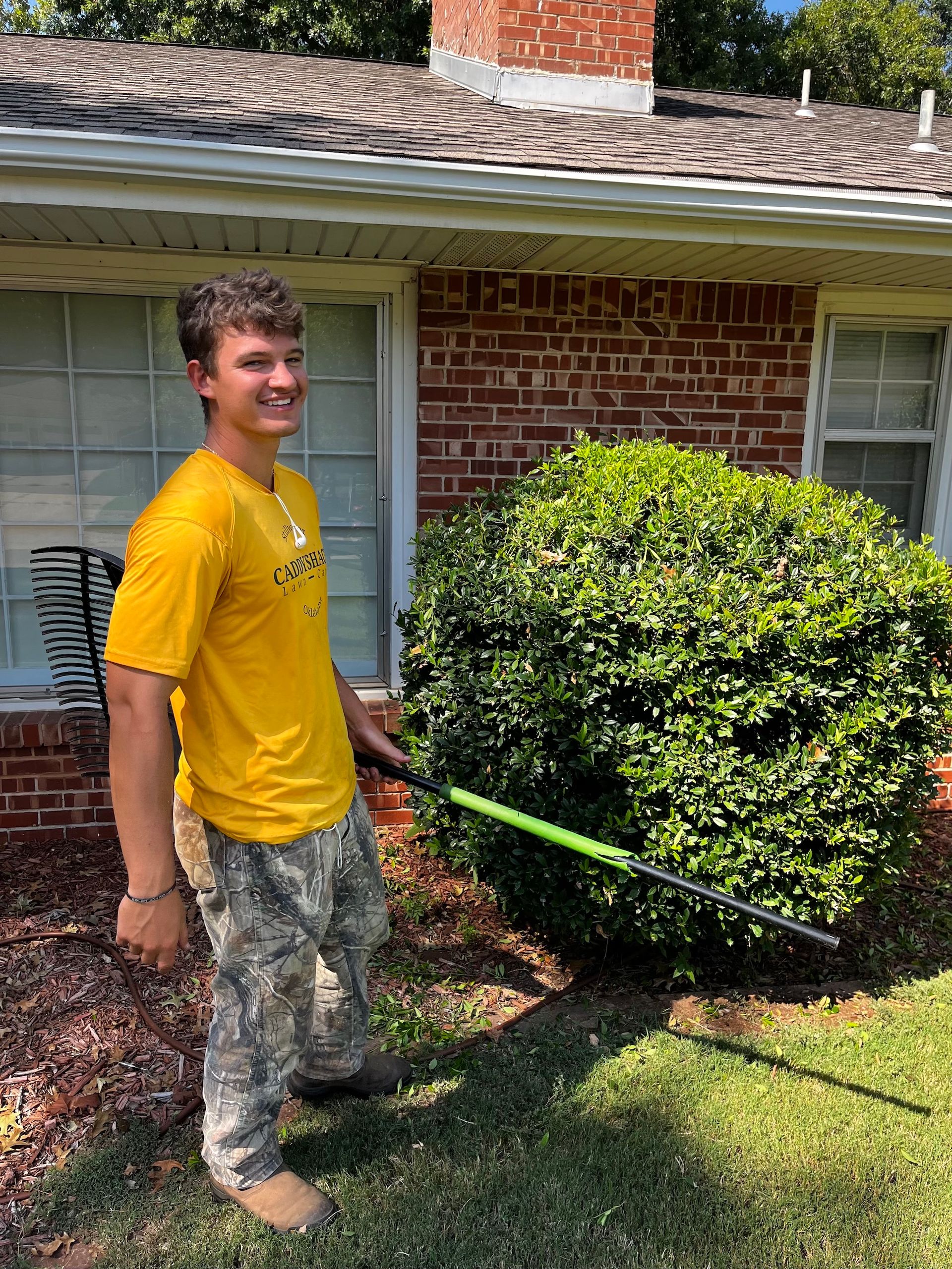 A young man in yellow shirt and camo pants smiling, holding a rake near a bush by a brick building.