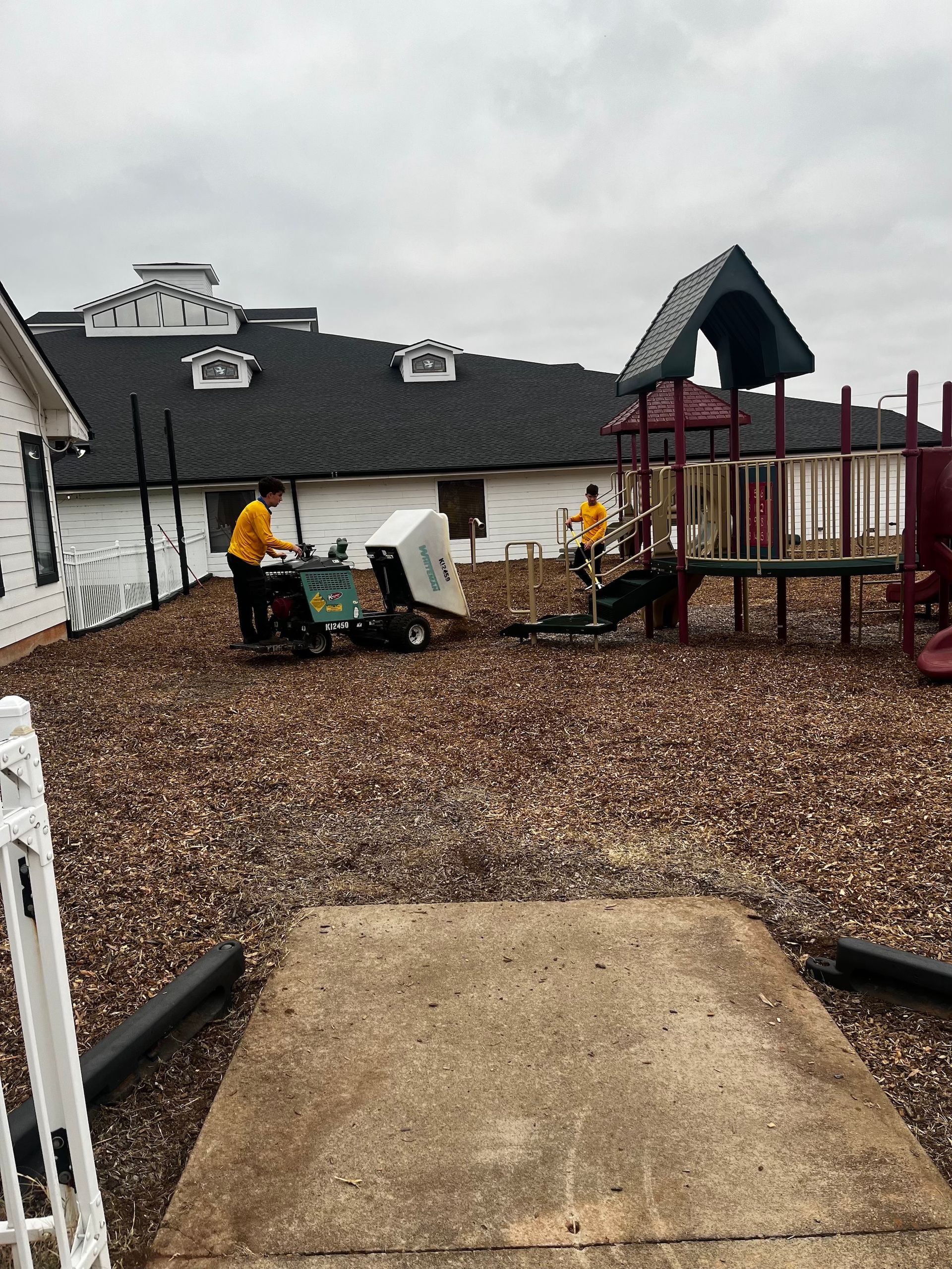Two workers moving a white object with a small machine on a playground covered in wood chips.