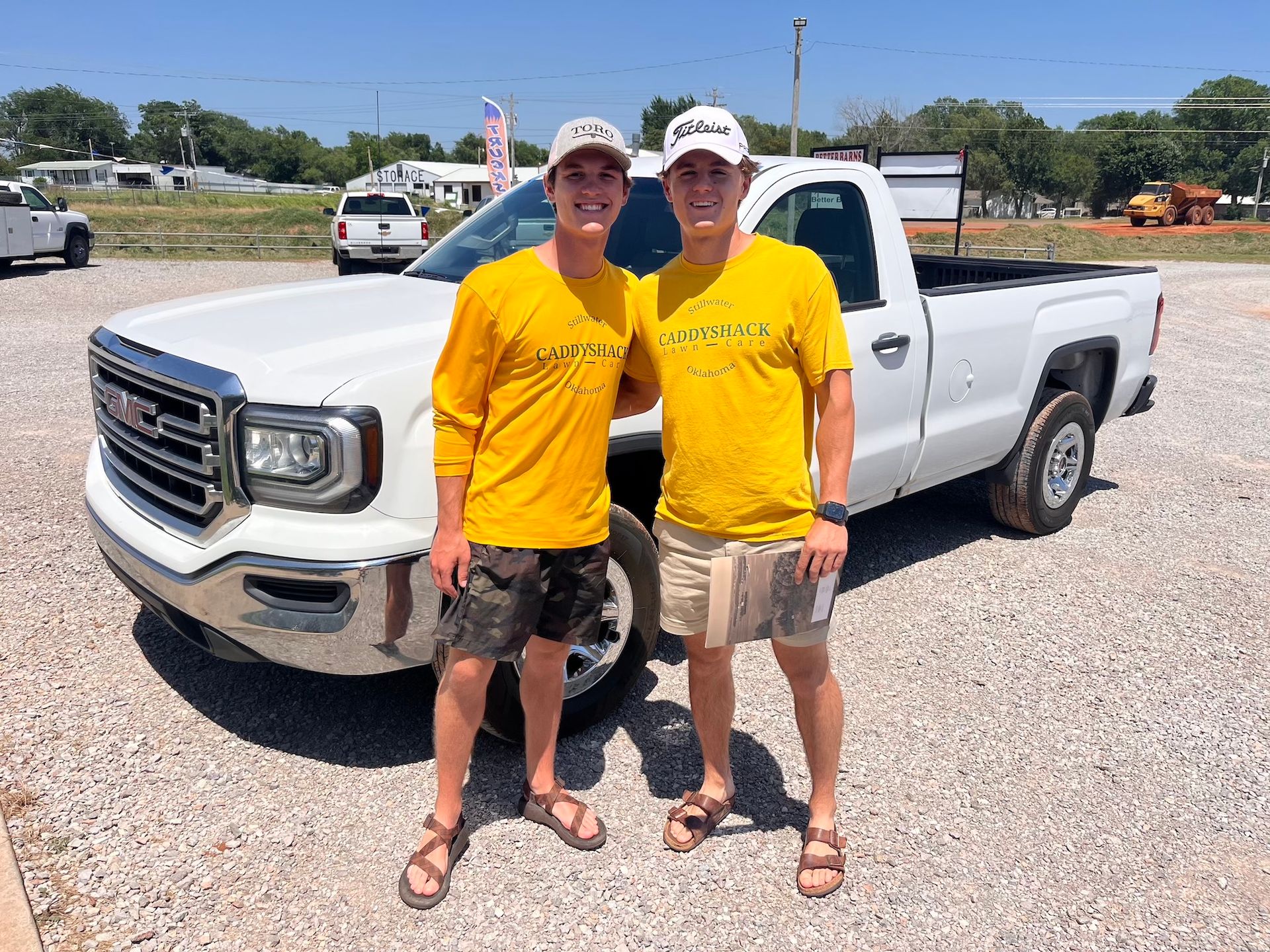 Two young men in yellow shirts stand by a white pickup truck on a sunny day.
