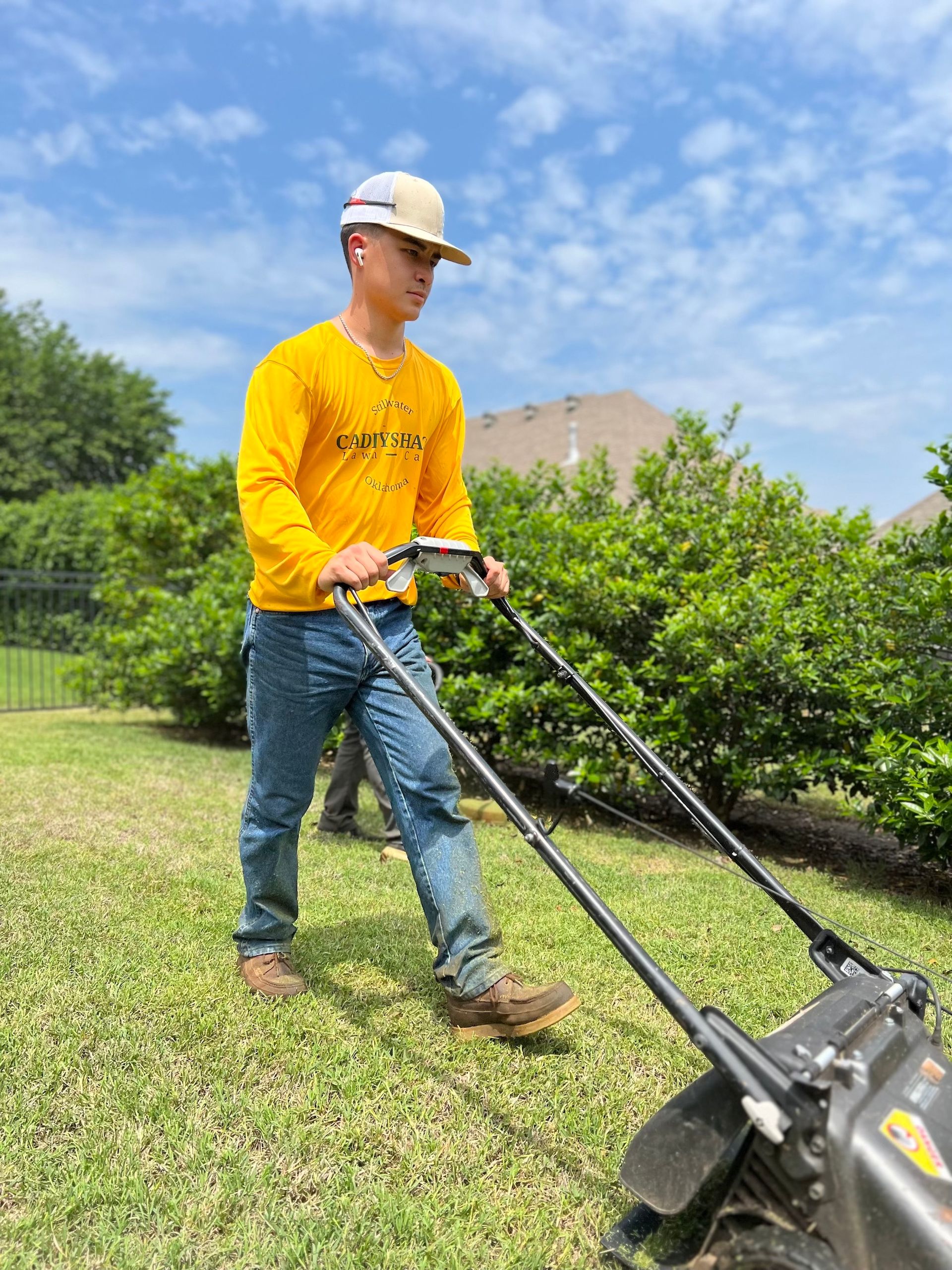 A young man mows a green lawn in a backyard, wearing a yellow shirt, jeans, and a hard hat.