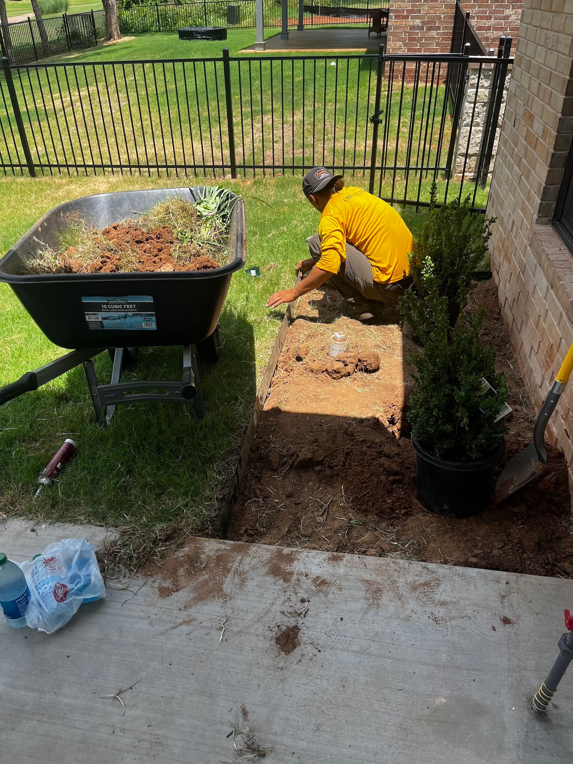 Person in yellow shirt planting by a building. Wheelbarrow with dirt and grass nearby.