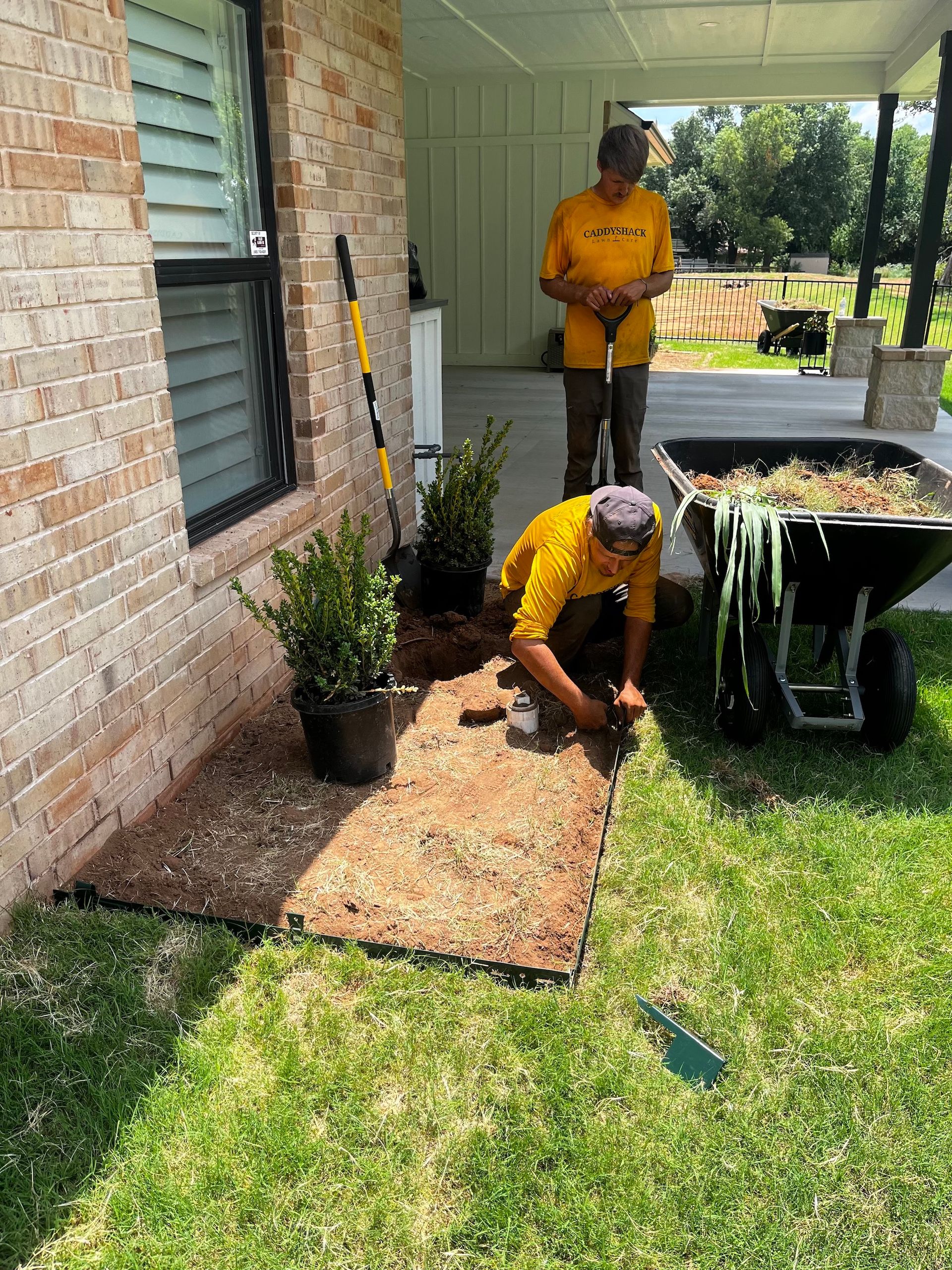 Two people planting near a brick house. One kneels, the other stands by a wheelbarrow filled with mulch.