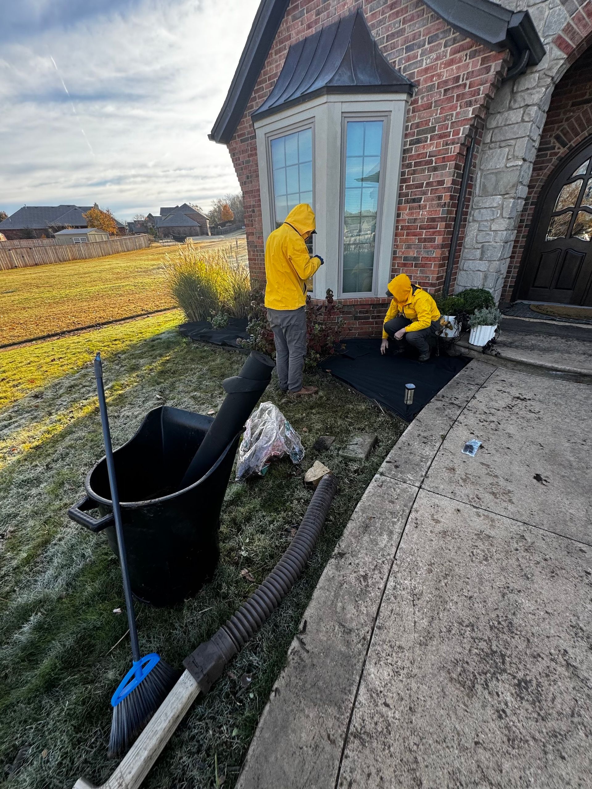 Two workers in yellow jackets install landscaping near a brick house, tools and debris are nearby.