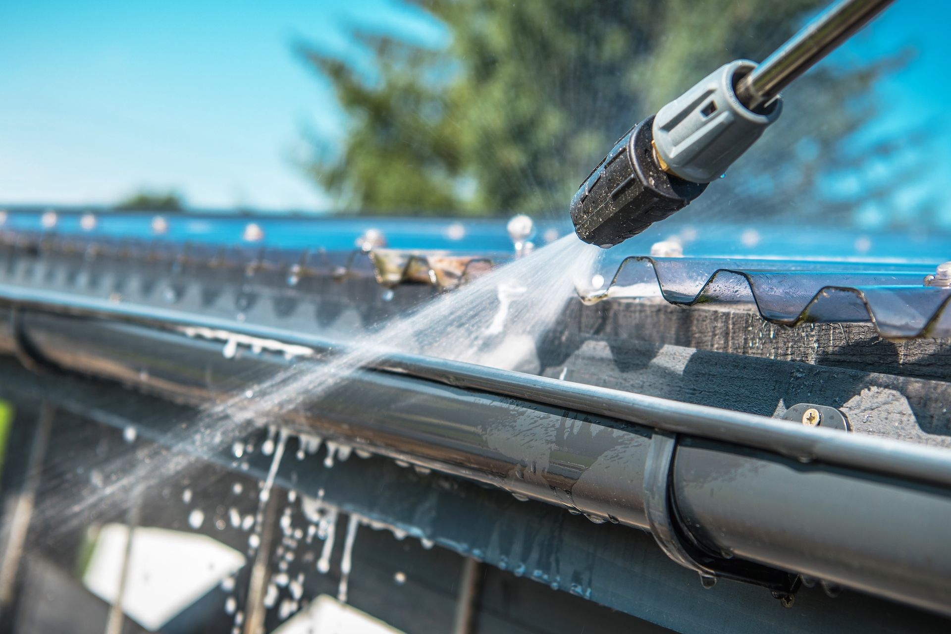 Pressure washing a gutter on a roof, water spraying, bright sunlight, outdoors.