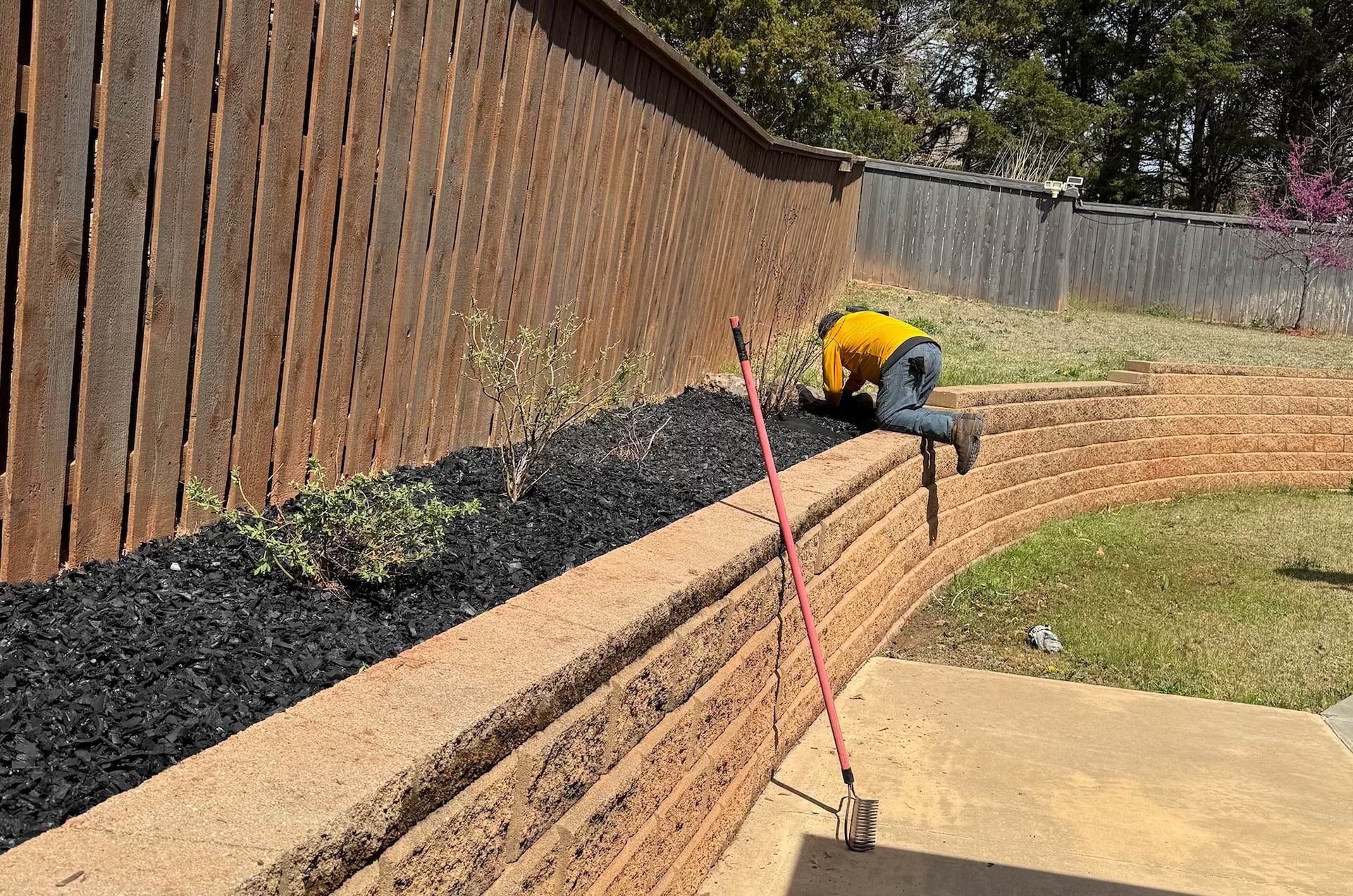A man is constructing a retaining wall in a landscaped area, surrounded by soil and plants.