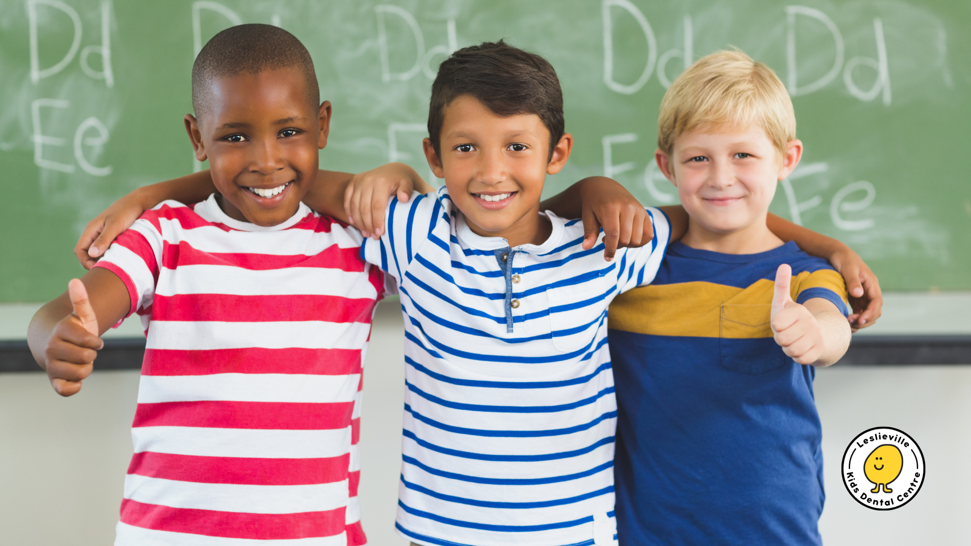 Three diverse children smiling, arms around each other, giving thumbs up in front of a chalkboard.