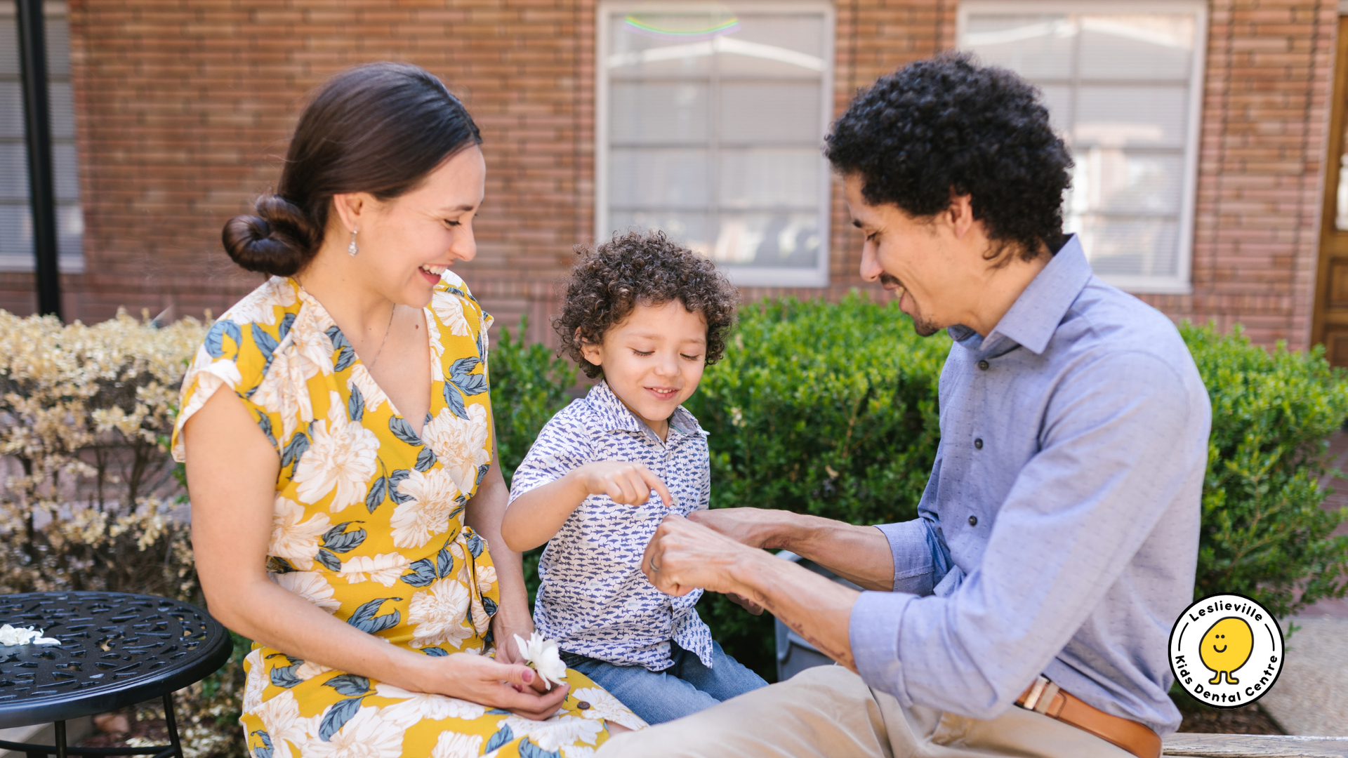 Family of three outside, smiling, playing with small object; sunny day.