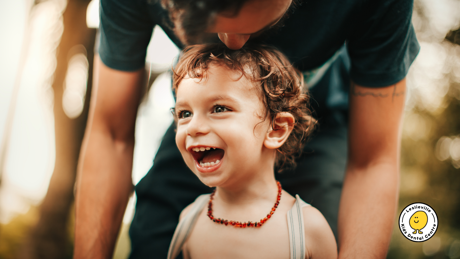 Person kissing a smiling child's head outdoors. Child wears amber necklace, suspenders. Sunlit trees in background.