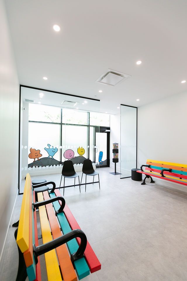 A brightly lit, minimalist waiting room featuring colorful benches, two black chairs, and whimsical wall decals by windows.