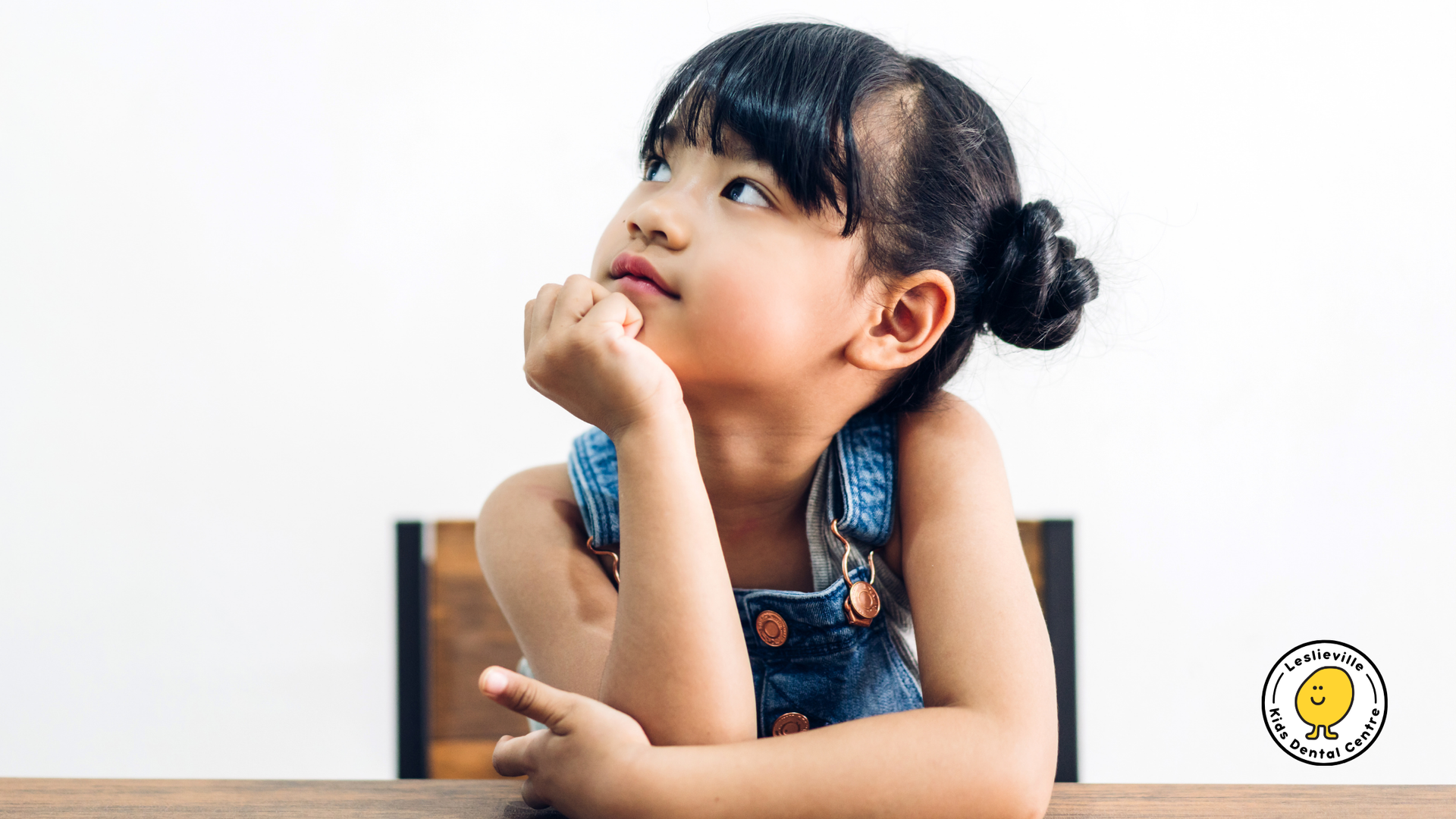 A little girl is sitting at a table with her hand on her chin and looking up.